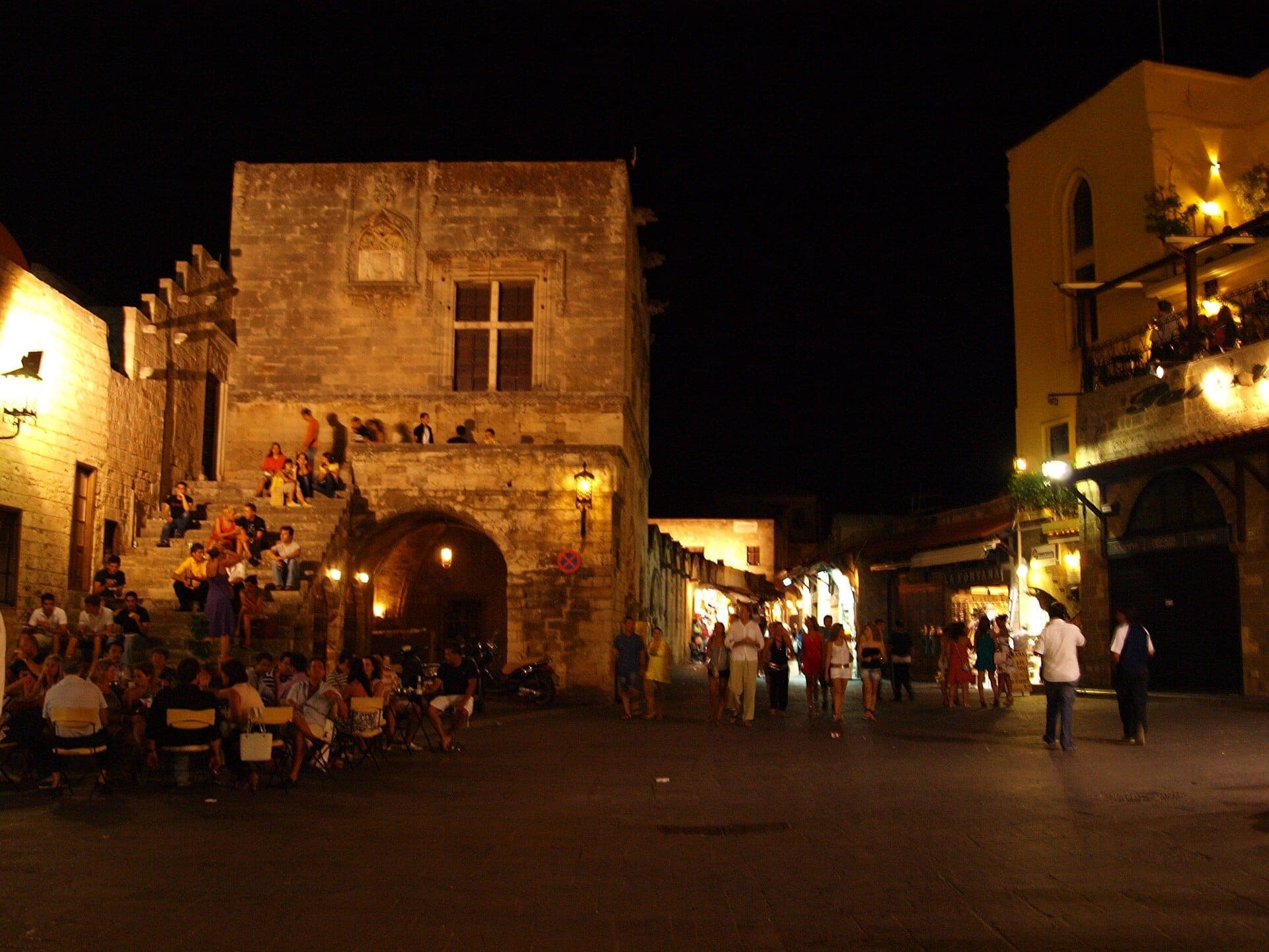 Des groupes de personnes rassemblées sur des marches en pierre et des tables en plein air dans la vieille ville médiévale de Rhodes la nuit, sous de chaudes lumières jaunes.