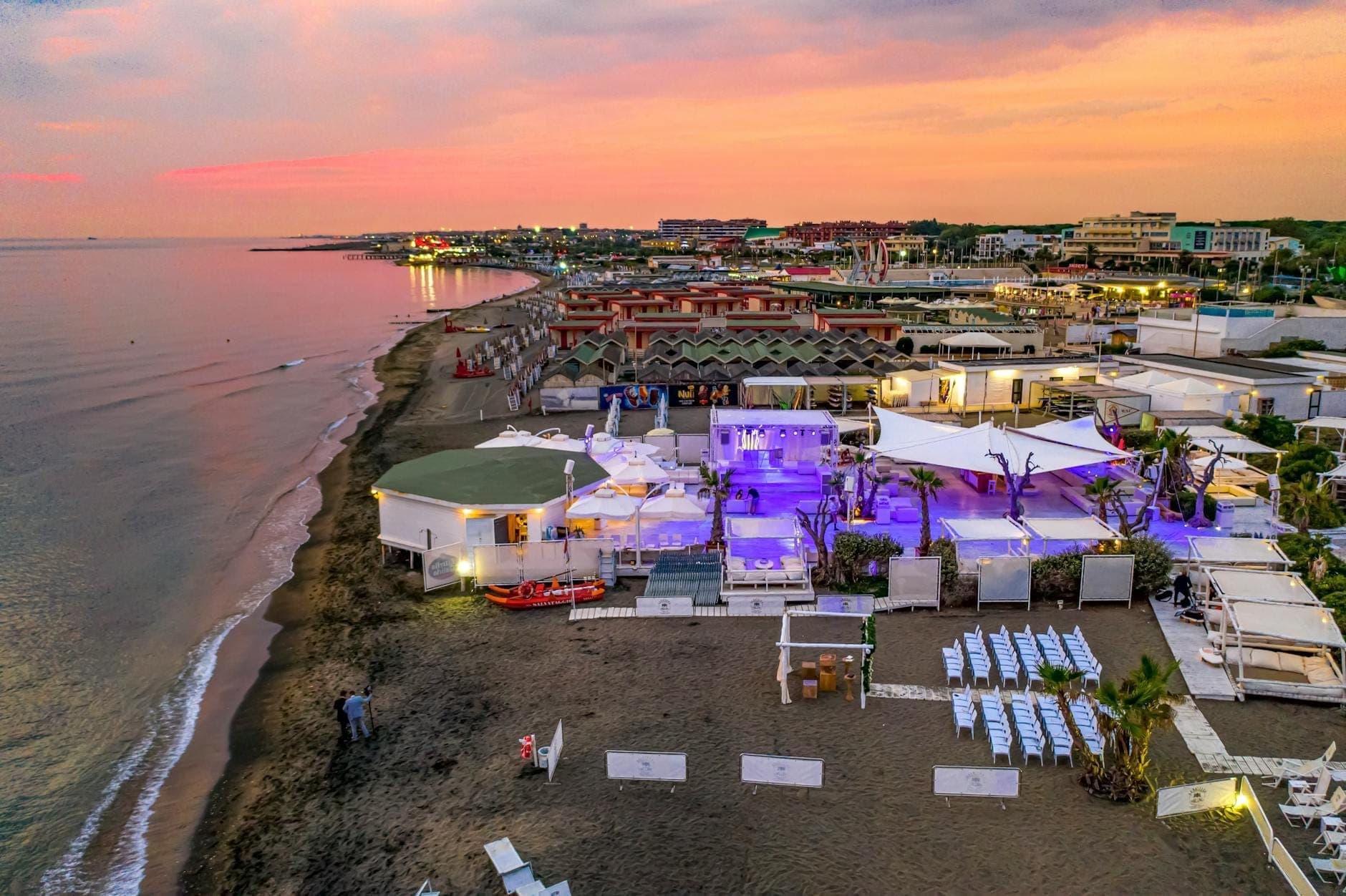 Tętniący życiem beach club o zachodzie słońca z fioletowym oświetleniem i stylowym wystrojem, usytuowany bezpośrednio na piasku w ruchliwej strefie nadmorskiej, przywołujący nocne życie Rodos.