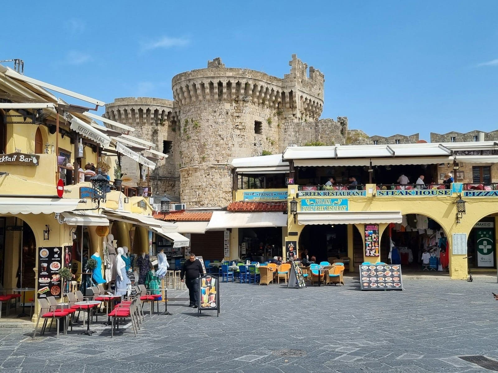 Place animée de la Vieille Ville de Rhodes avec des cafés aux terrasses colorées et les imposantes fortifications médiévales en pierre se dressant en arrière-plan, par une belle journée ensoleillée.