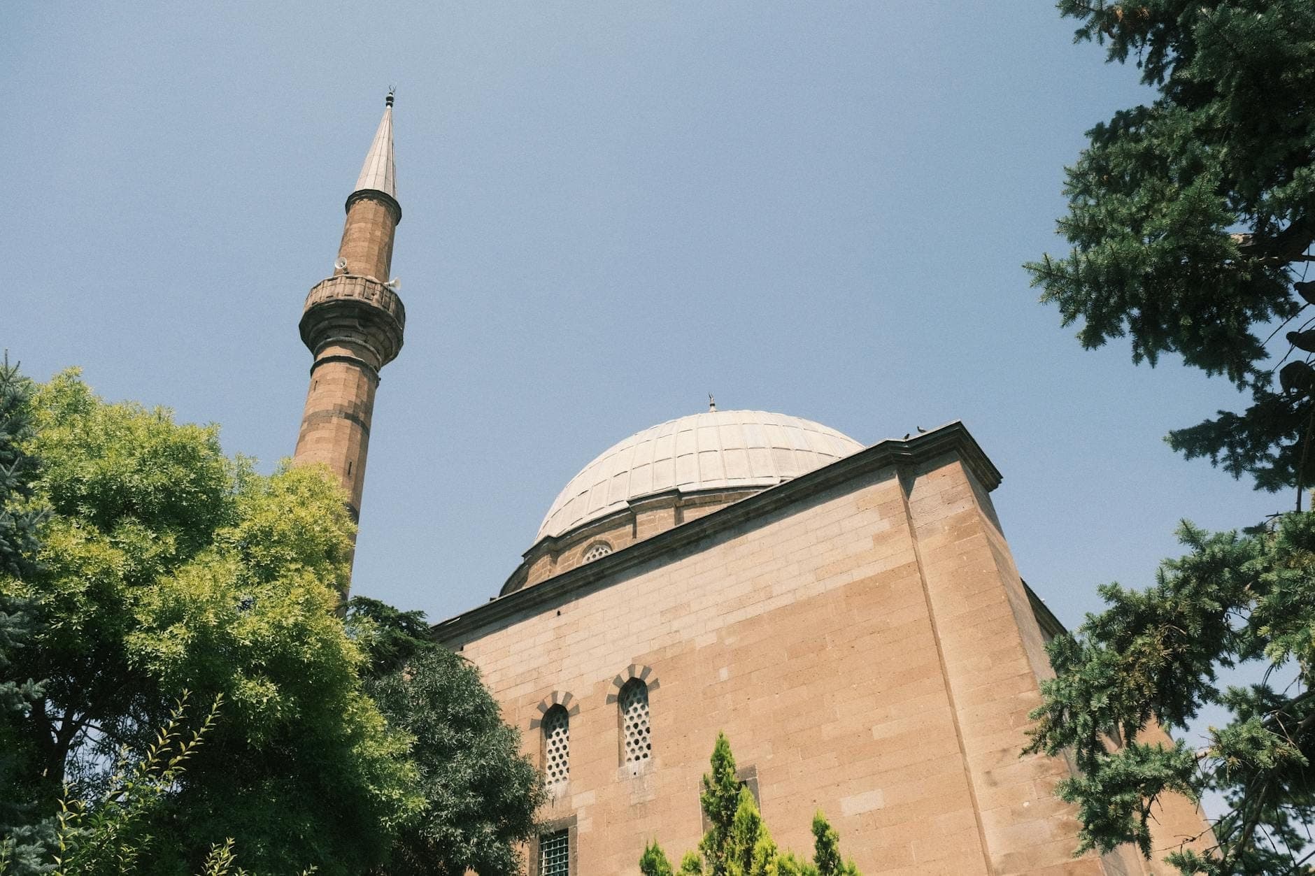 A stone mosque with a single minaret rises behind green trees under a clear sky, showing typical Ottoman architecture.