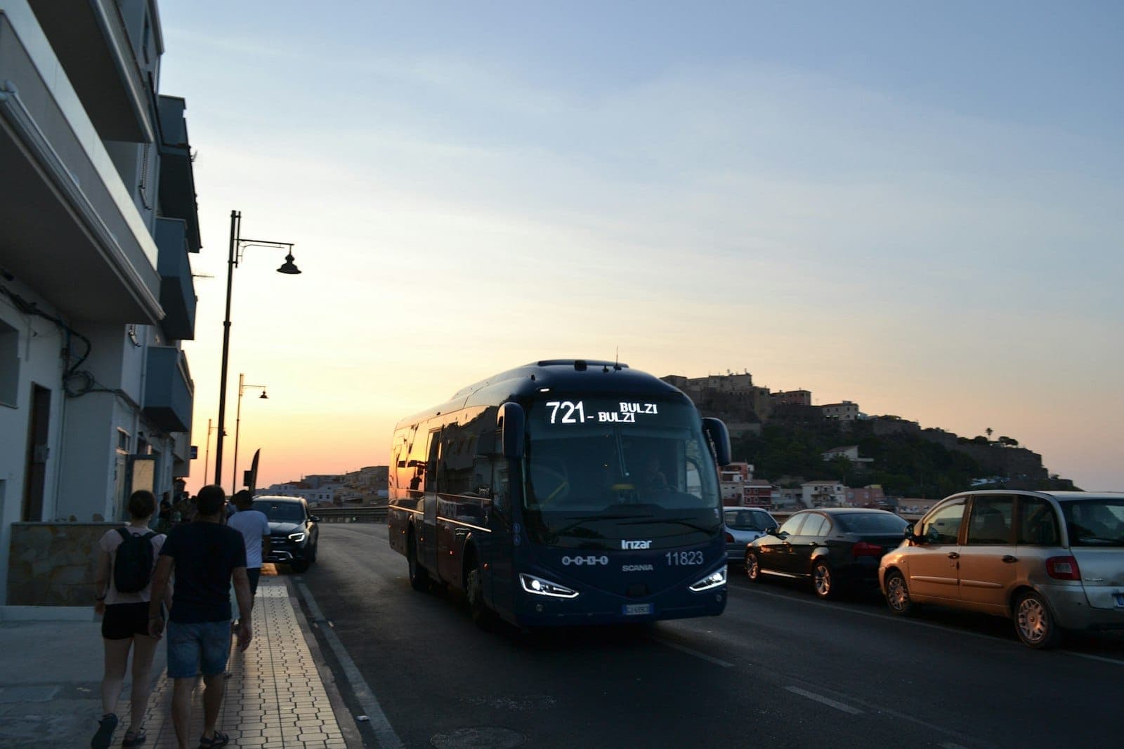 Bus public roulant le long d'une route en bord de mer à Rhodes au coucher du soleil, avec des passants sur le trottoir et un monument sur une colline en arrière-plan.