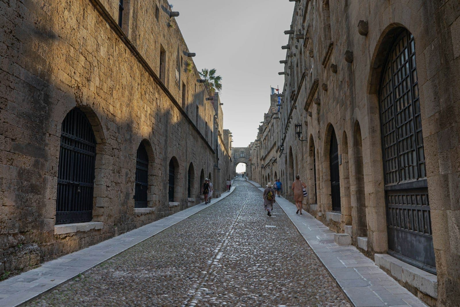 Rue pavée ensoleillée bordée de bâtiments médiévaux en pierre dans la vieille ville de Rhodes, avec des promeneurs au loin.