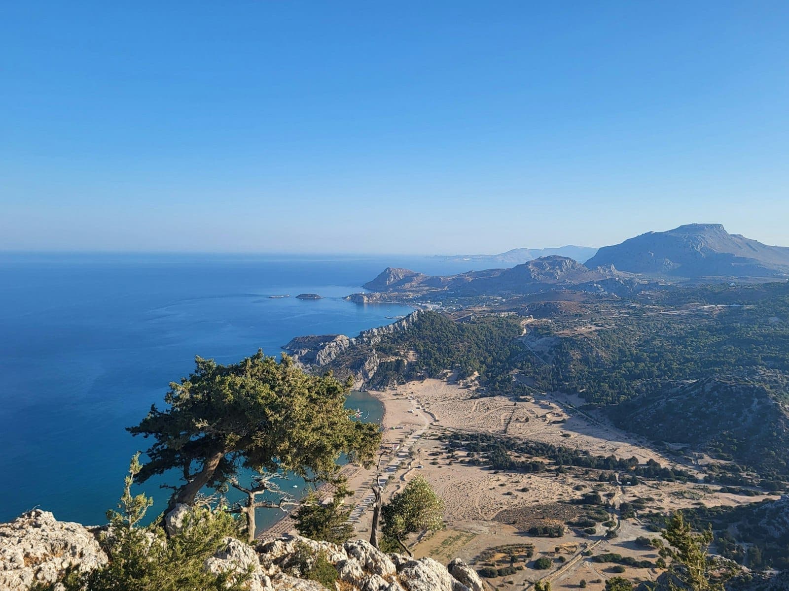 Vue panoramique spectaculaire depuis un belvédère dominant un arbre, une plage de sable, une baie turquoise et des montagnes sauvages sur l'île de Rhodes sous un ciel bleu immaculé.