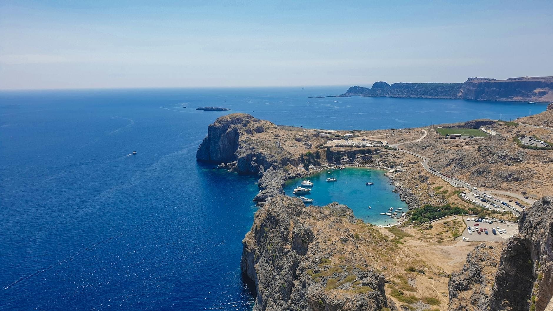 A stunning aerial view of a rocky cove with turquoise water in Rhodes, boats anchored in the bay, and rugged coastline meeting the deep blue Aegean Sea.