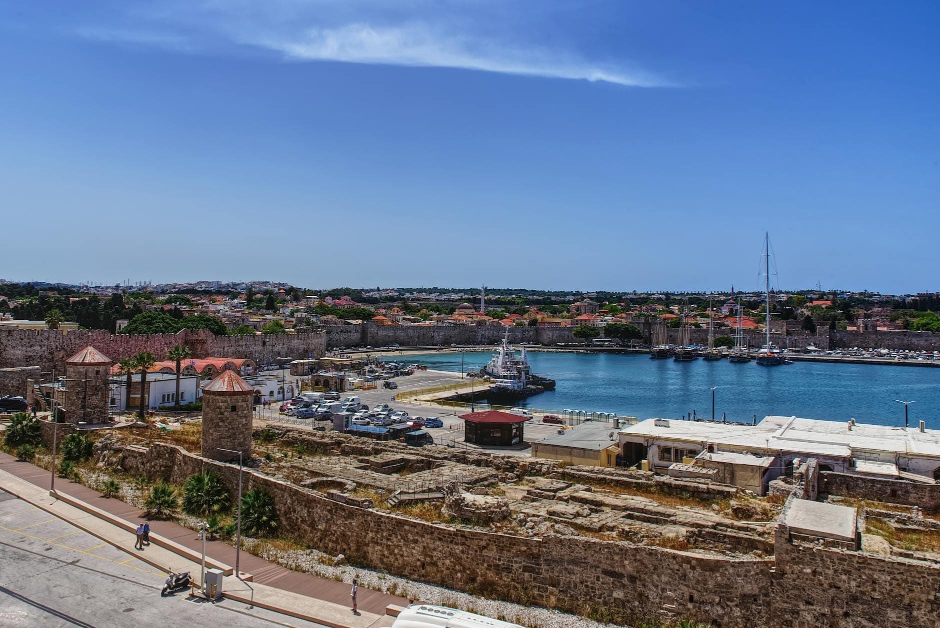 Vue panoramique du port commercial de Rhodes avec les remparts médiévaux en pierre, des bateaux amarrés et la ville de Rhodes en arrière-plan sous un ciel bleu dégagé.