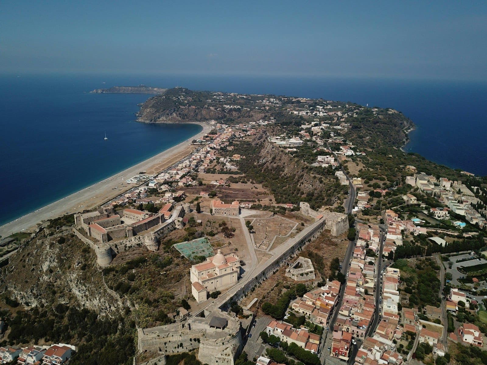 Vue aérienne de la péninsule de la ville de Rhodes, avec une plage de sable, des remparts historiques et une mer bleue entourée de bâtiments.