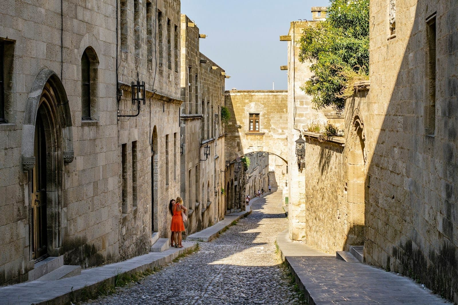 Une rue médiévale en pierre ensoleillée dans la vieille ville de Rhodes avec des bâtiments historiques, des portes en arc et un couple se promenant sous une arche.
