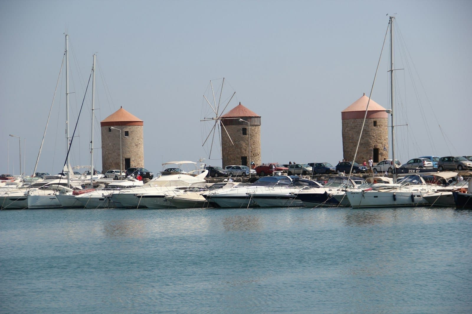 Trois moulins à vent médiévaux en pierre sur le port de Mandraki à Rhodes avec des yachts amarrés devant sous un ciel dégagé.