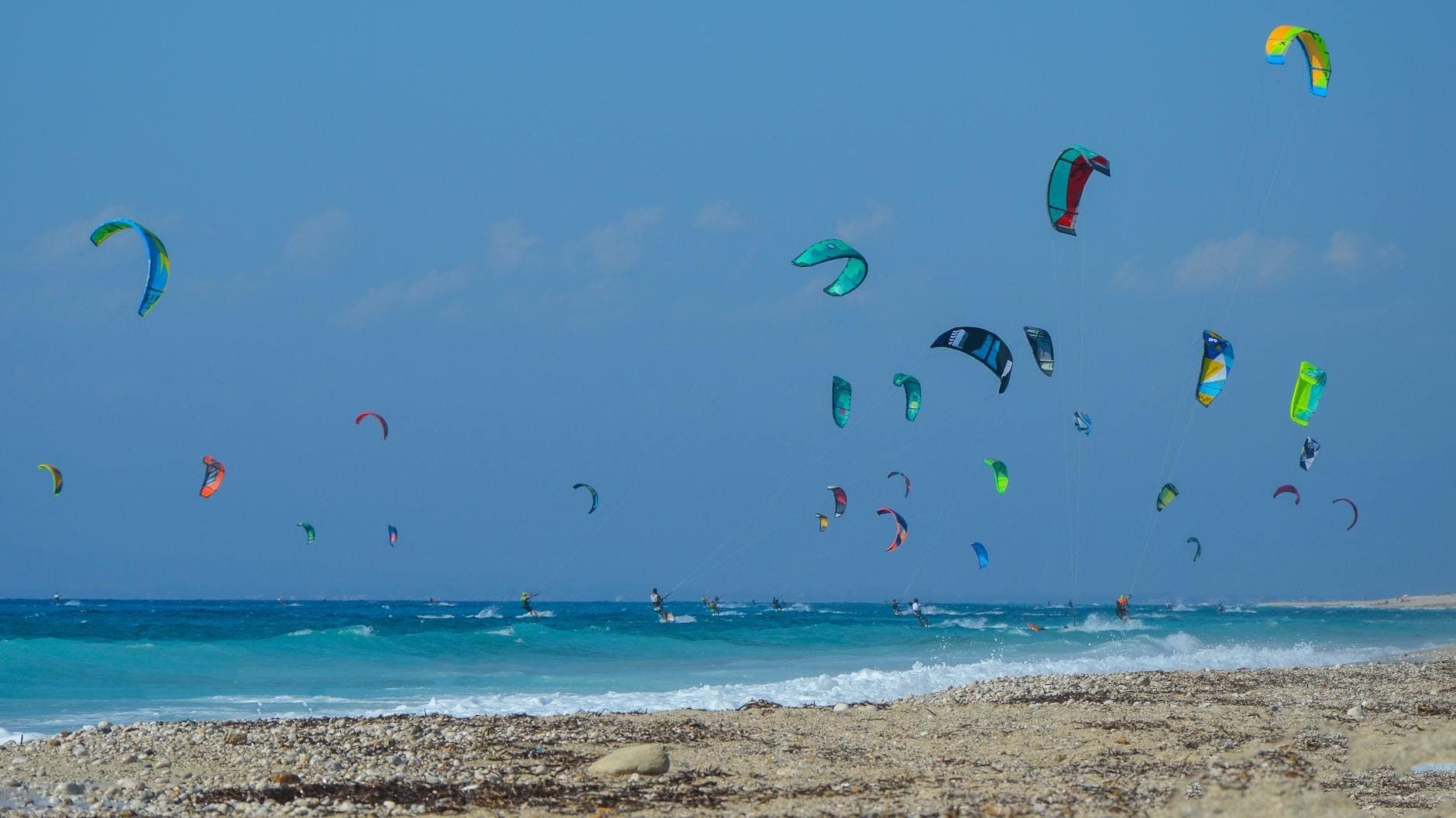 Dziesiątki kolorowych latawców kitesurfingowych zdobią błękitne niebo nad turkusowymi falami i kamienistą plażą, oddając atmosferę sportów wodnych na Rodos.