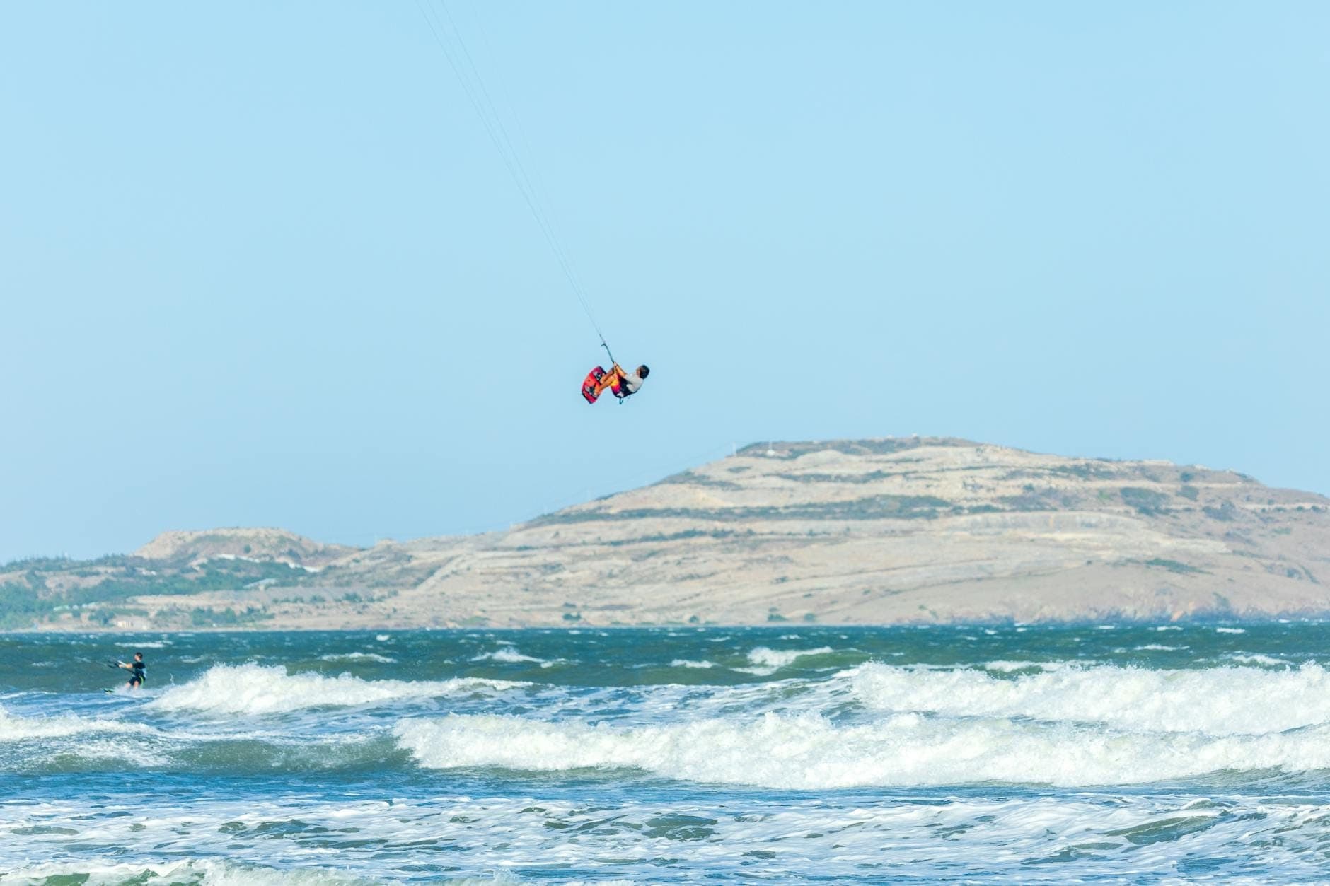 Kitesurfer jadący na dużych falach przy wietrznym wybrzeżu z suchym, pagórkowatym krajobrazem Rodos w tle, ukazujący idealne warunki do sportów wietrznych.