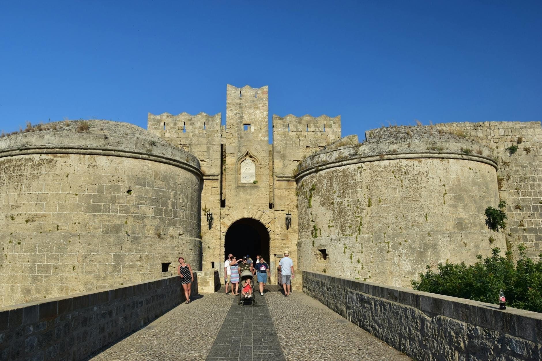 Des familles avec enfants franchissent le pont de pierre menant à l'entrée d'un château médiéval sous un ciel bleu éclatant, à Rhodes, en Grèce.
