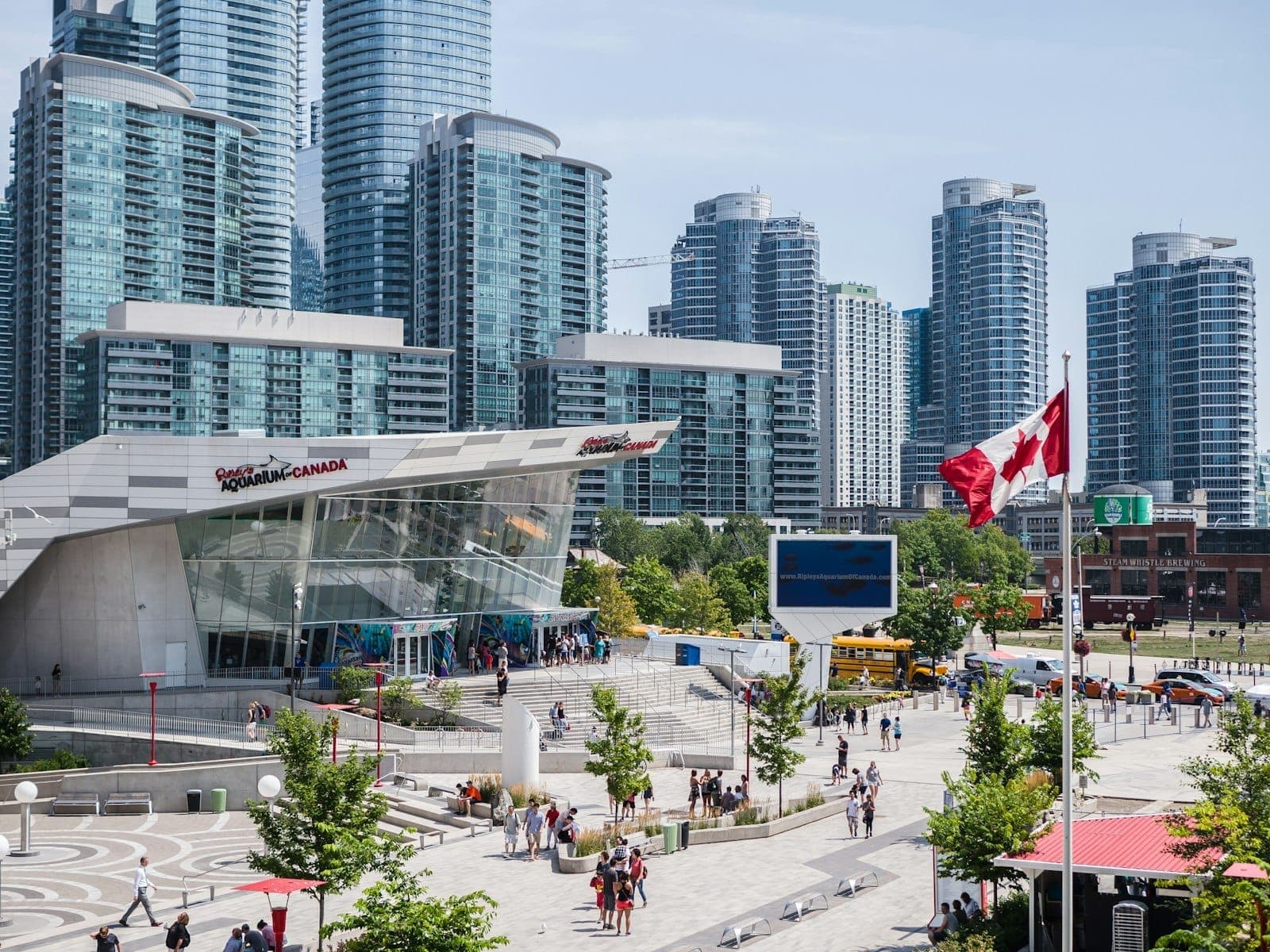 Ingresso del Ripley's Aquarium of Canada con visitatori all'esterno, moderno edificio in vetro, bandiera canadese e grattacieli di Toronto sullo sfondo in una giornata di sole.