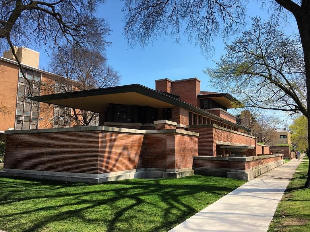 Robie House en Hyde Park, un llamativo edificio de ladrillo bajo con líneas horizontales, rodeado de césped verde y árboles primaverales bajo un cielo azul despejado.