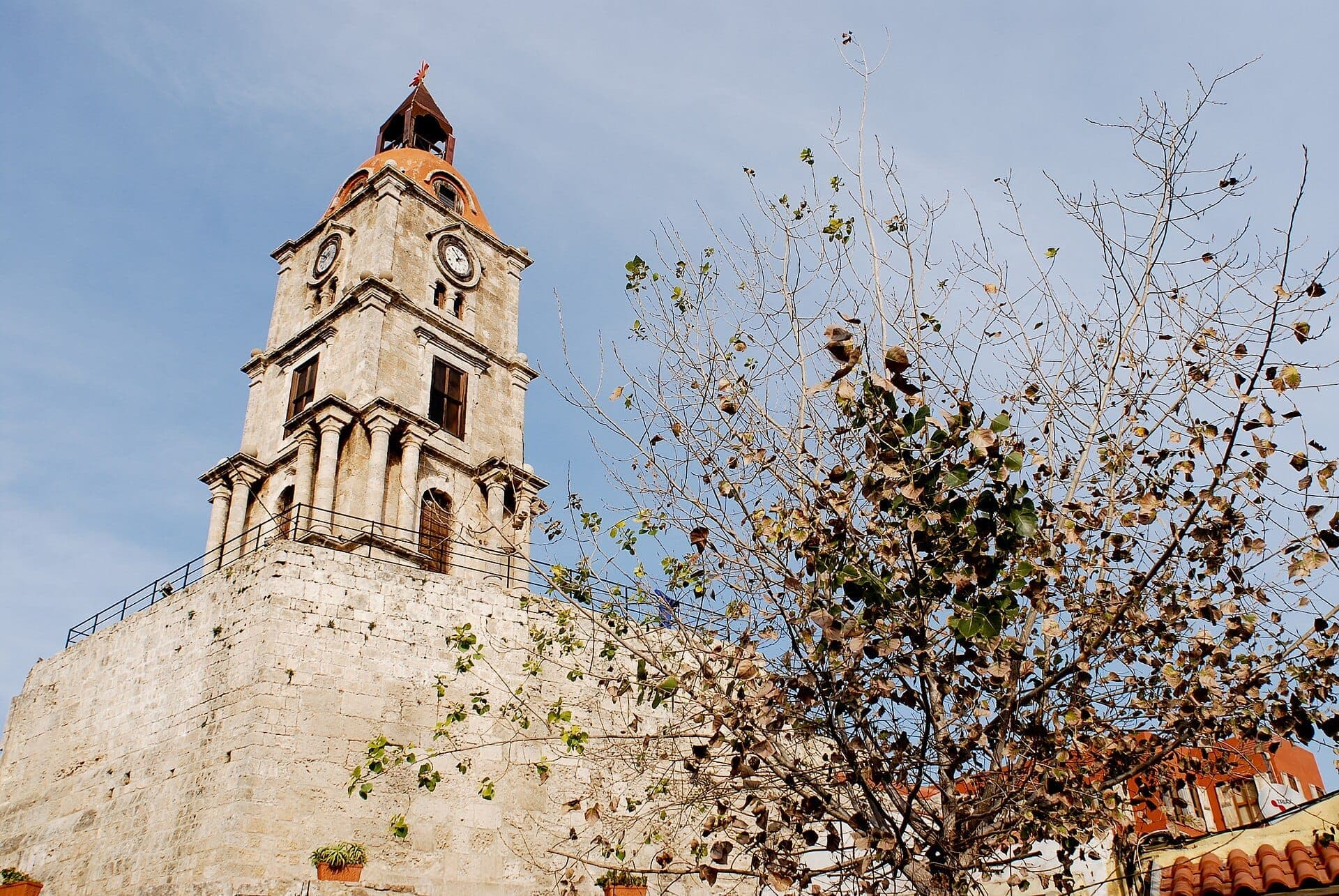 La Tour de l'Horloge Roloi dans la vieille ville de Rhodes s'élève au-dessus de murs en pierre près d'un arbre feuillu, sous un ciel bleu dégagé.