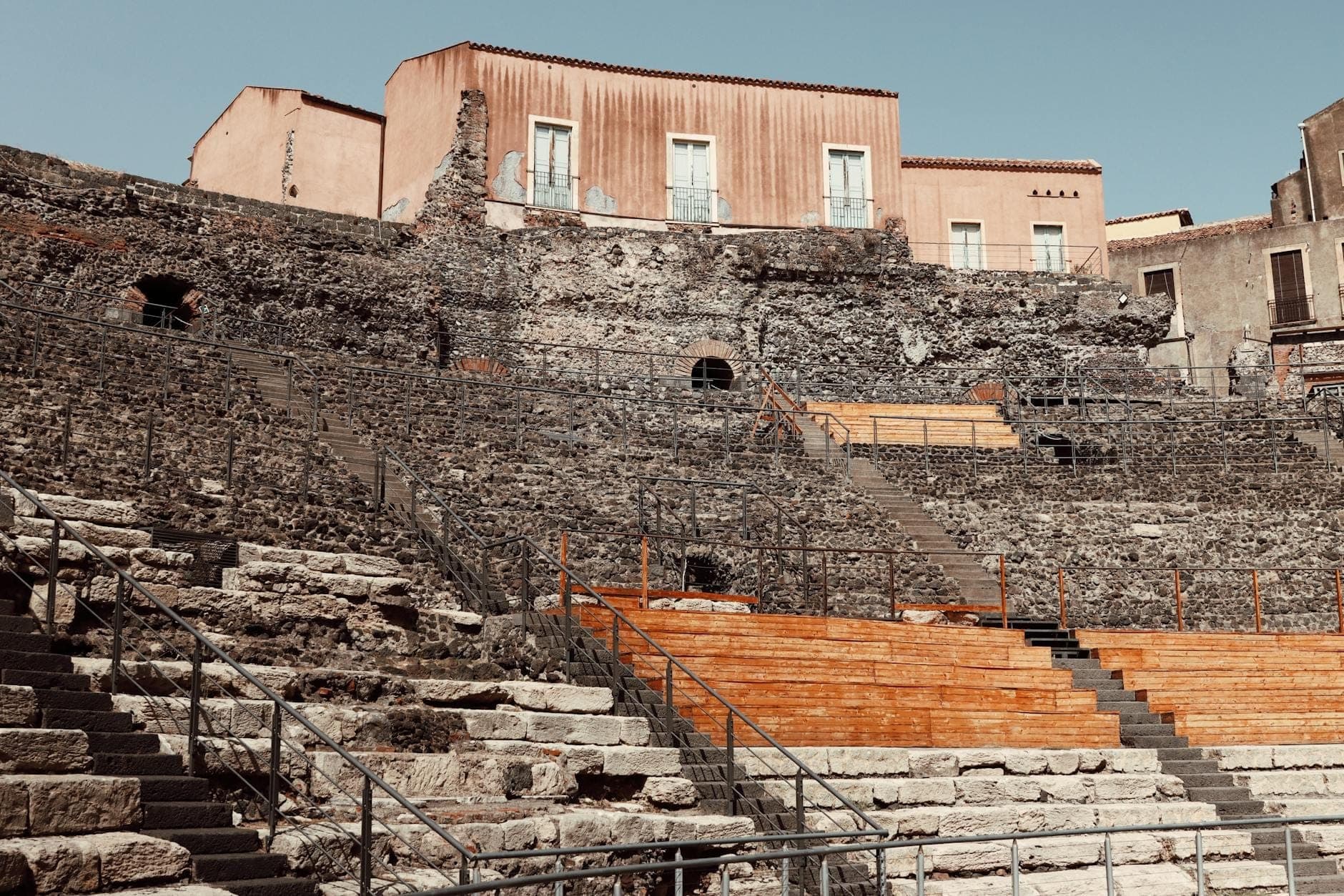 Tempat duduk batu bertingkat Amfiteater Romawi Catania terlihat di bawah gedung-gedung pusat kota di bawah langit cerah, memperlihatkan reruntuhan kuno dan kontras dengan dunia modern.