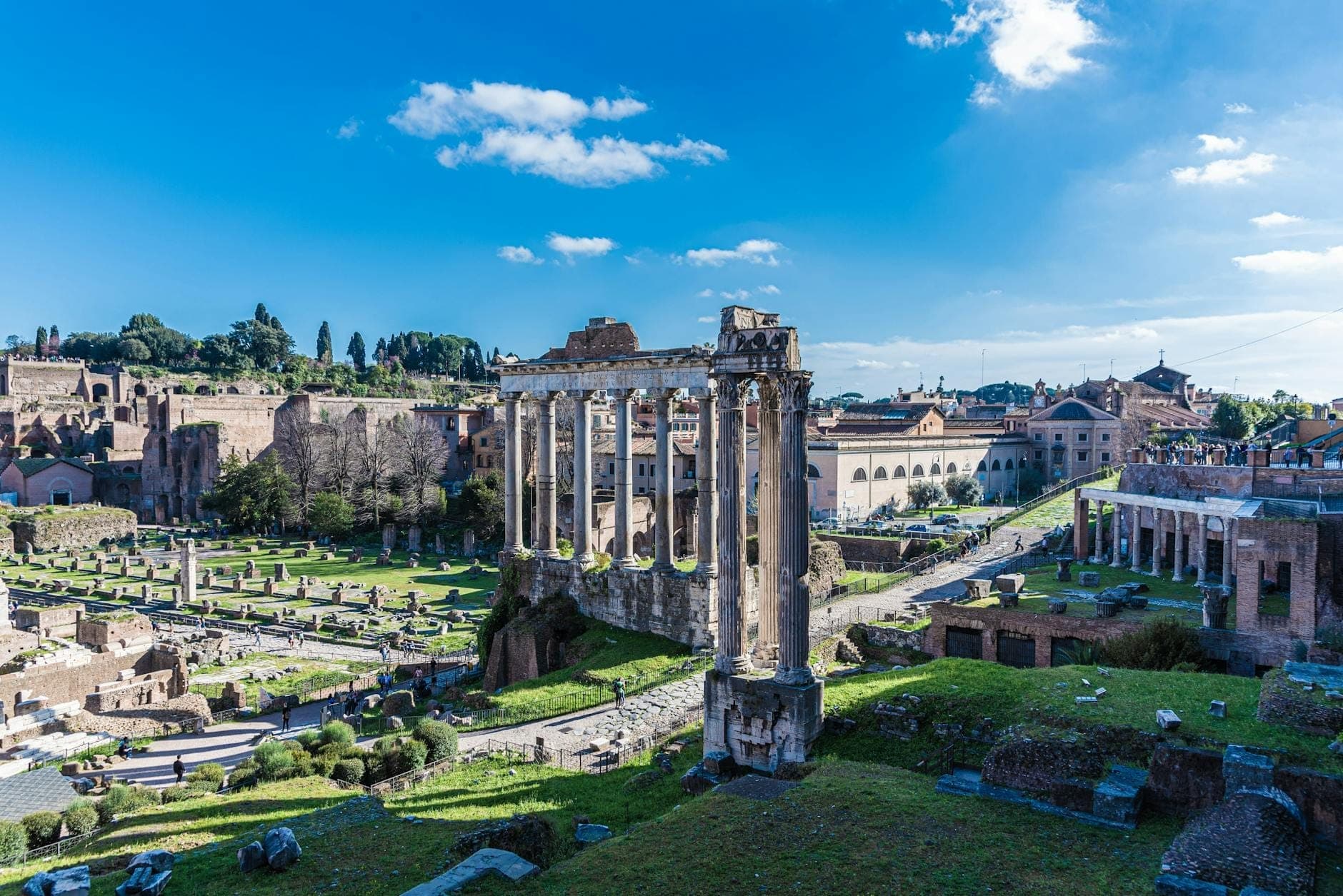 Vista panoramica del Foro Romano sotto un cielo azzurro intenso, con le rovine di antichi templi, basiliche e colonne circondate da prati verdi.