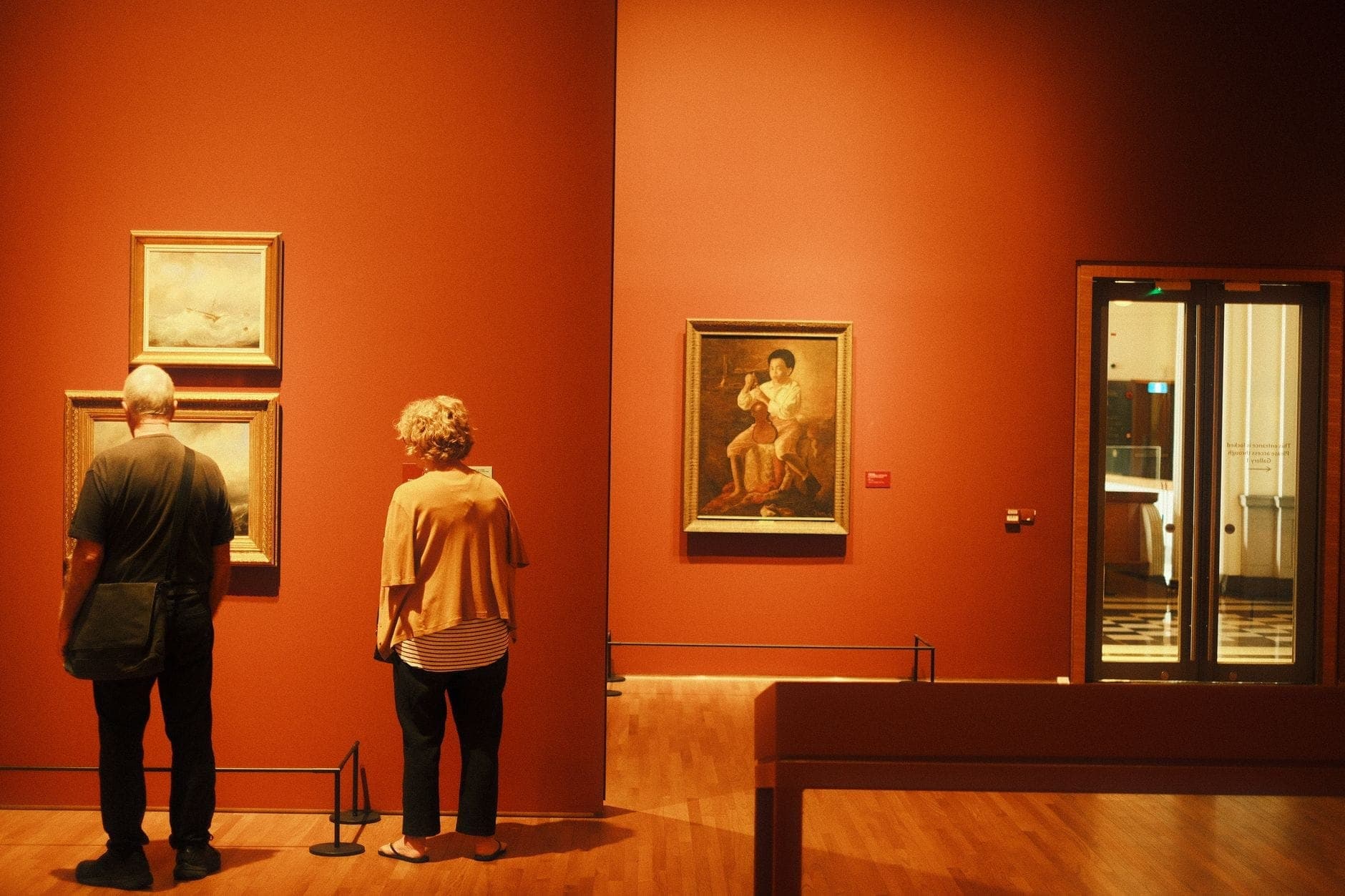 Un couple se tient dans une galerie de musée d'art chaleureusement éclairée, admirant des peintures encadrées exposées sur des murs rouge profond.