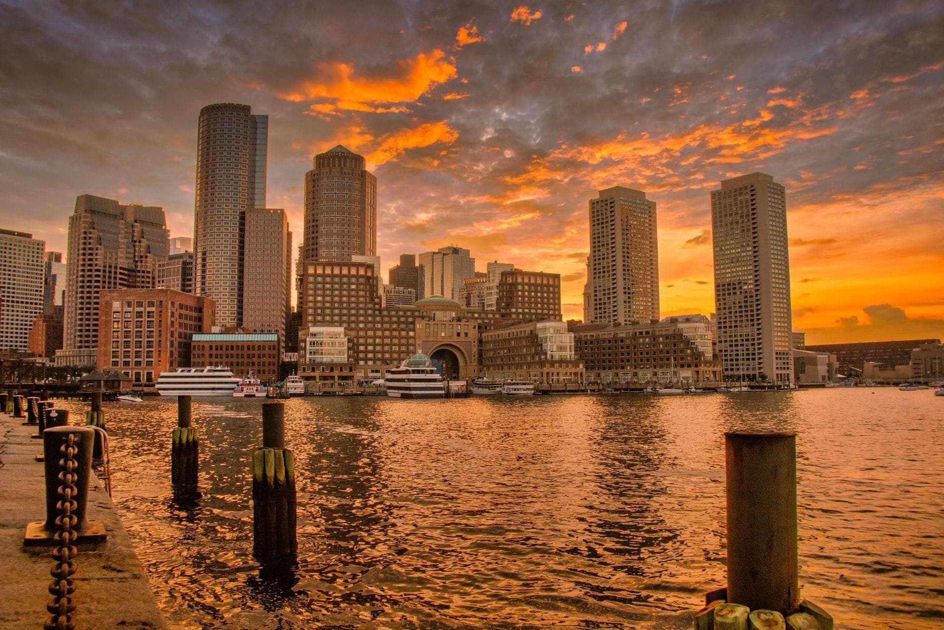 Coucher de soleil spectaculaire sur le skyline de Boston reflété dans les eaux du port, avec des nuages orangés et des bateaux amarrés le long des quais.