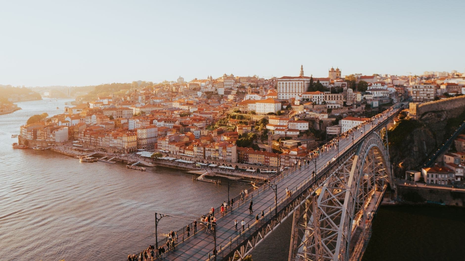 Aerial na tingin ng Tulay na Dom Luís I sa ibabaw ng Ilog Douro sa pagsalubin ng araw, na may mga taong naglalakad sa tulay at makasaysayang tanawin ng lungsod ng Porto sa likod.