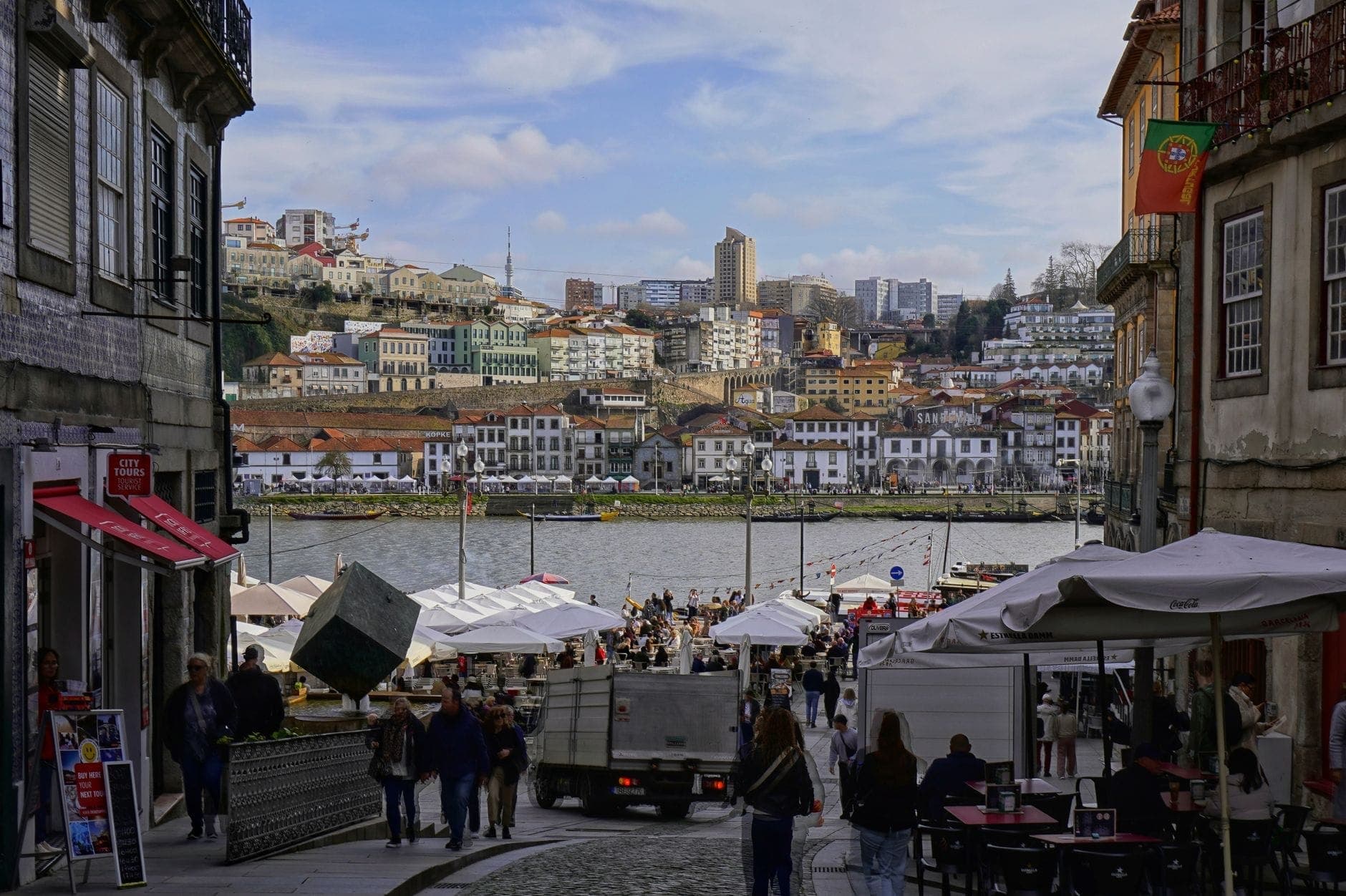 Tanawin ng kalye patungo sa dalampasigan ng ilog Douro sa Porto na may makulay na mga gusali, mga outdoor na café, at mga taong naglalakad, na kumukuha ng masaya ngunit malapit na kapaligiran ng lungsod.