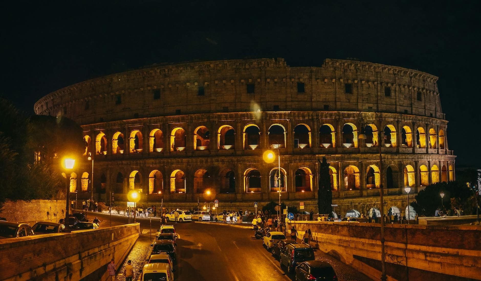 Góc nhìn rộng của Đấu trường La Mã Colosseum ở Rome được chiếu sáng vào ban đêm, với những vòm cung rực sáng và xe ô tô đậu trên đường phía trước.