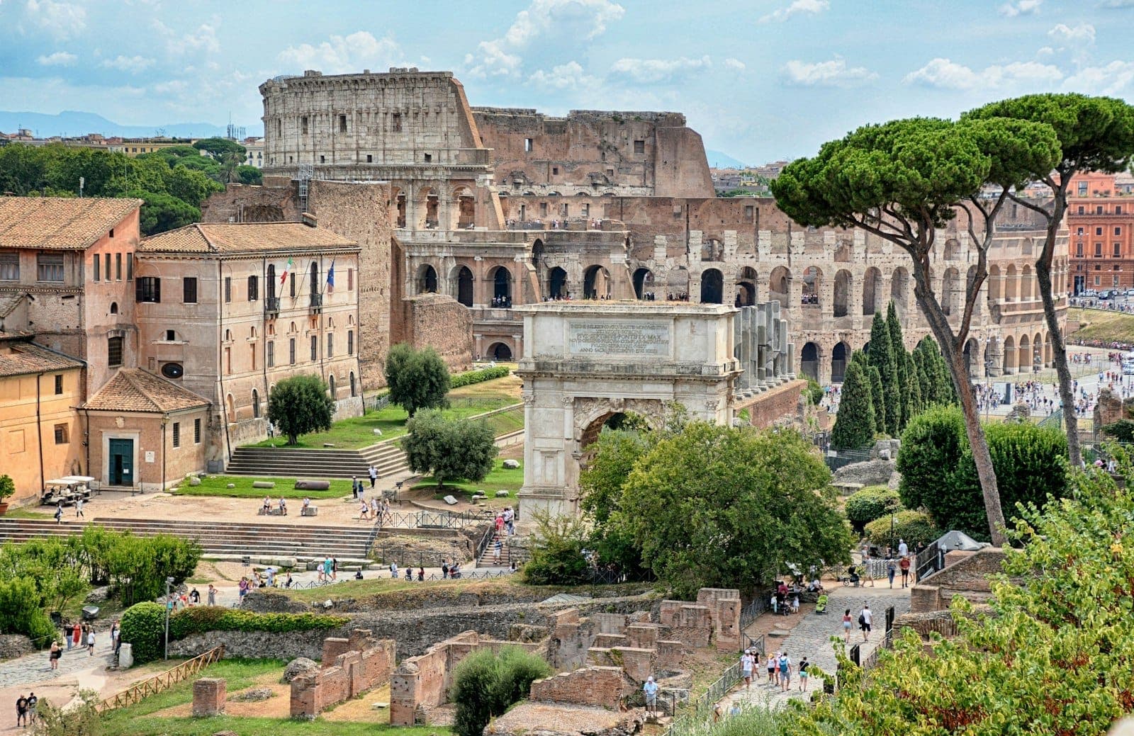 Tingin sa Colosseum at Arch of Constantine na napapaligiran ng mga sinaunang guho at mga puno ng payong sa maliwanag na langit, nagpapaalala ng mayamang kasaysayan ng Roma.