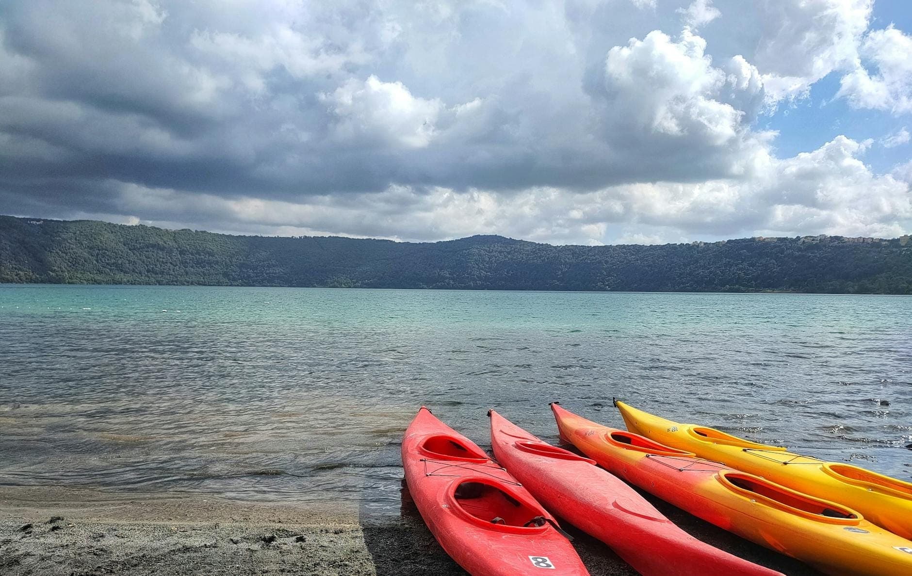 Bờ của một hồ núi lửa với nhiều chiếc thuyền kayak đầy màu sắc trên cát, những ngọn đồi có rừng cây ở phía xa và những đám mây ấn tượng phía trên.