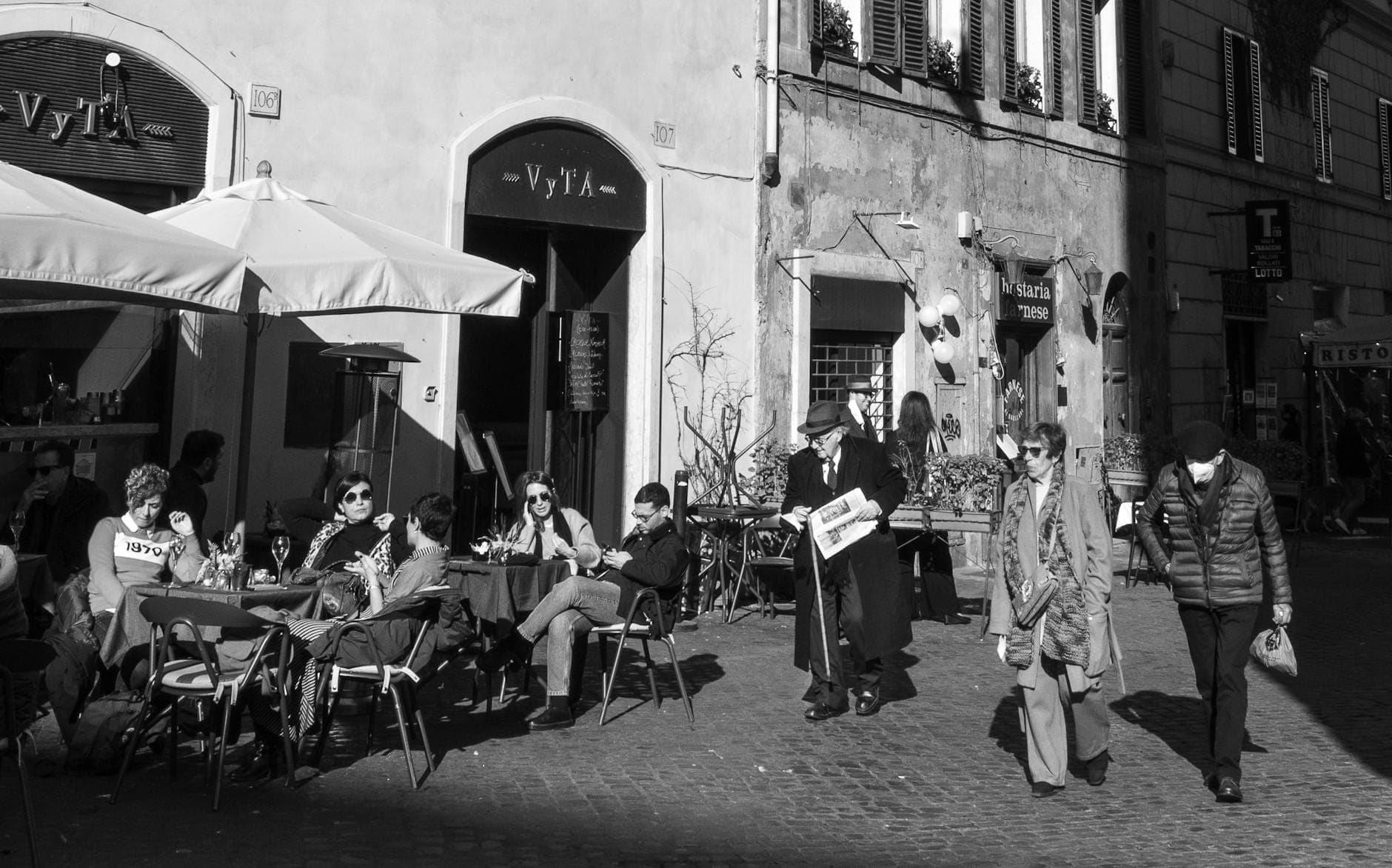 Foto em preto e branco de pessoas jantando em mesas ao ar livre perto de edifícios históricos, evocando o charme do Ghetto Judeu de Roma.