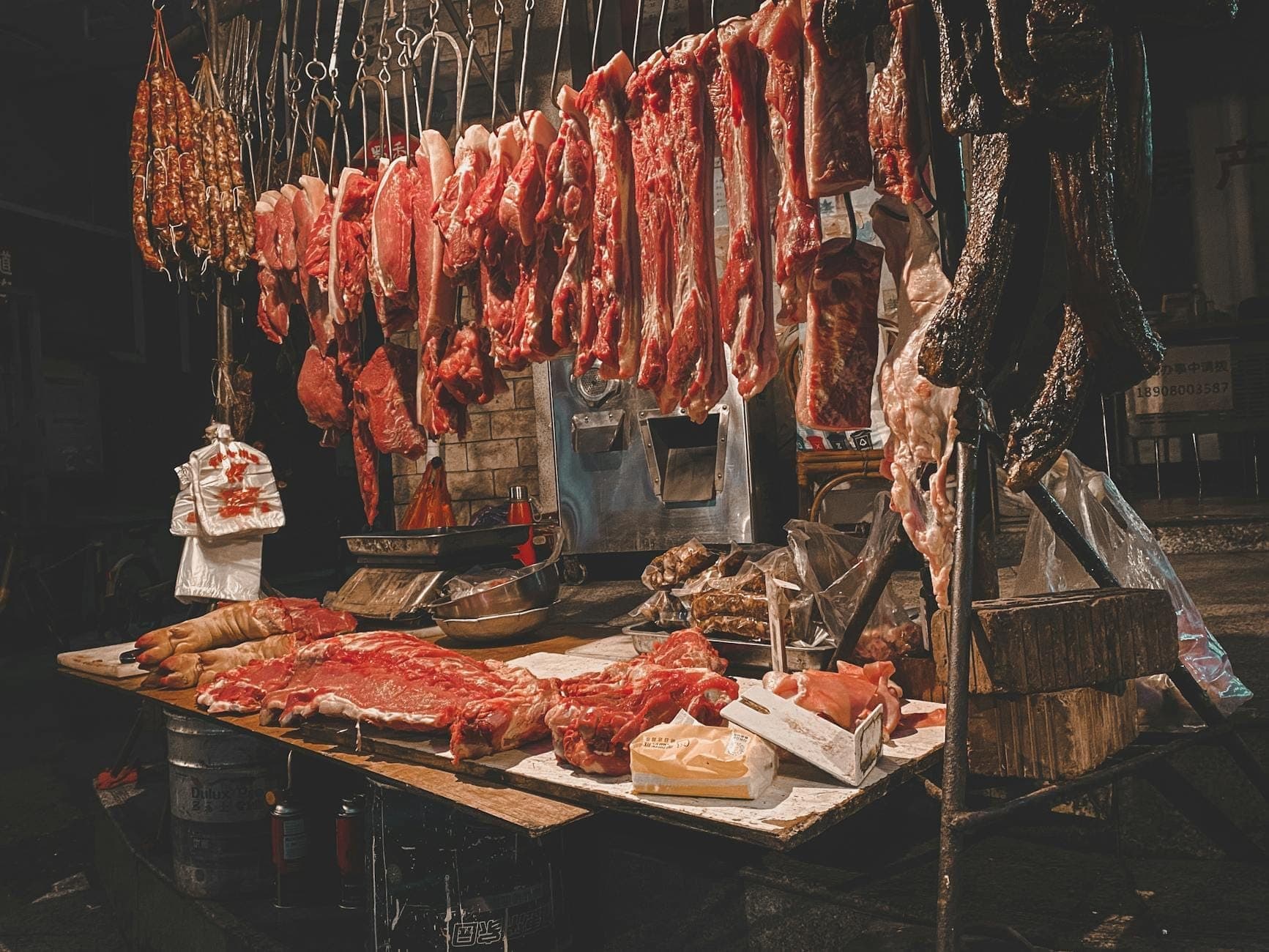 Banca de açougue com vários cortes de carne e miúdos pendurados e expostos, iluminados em uma atmosfera de mercado.