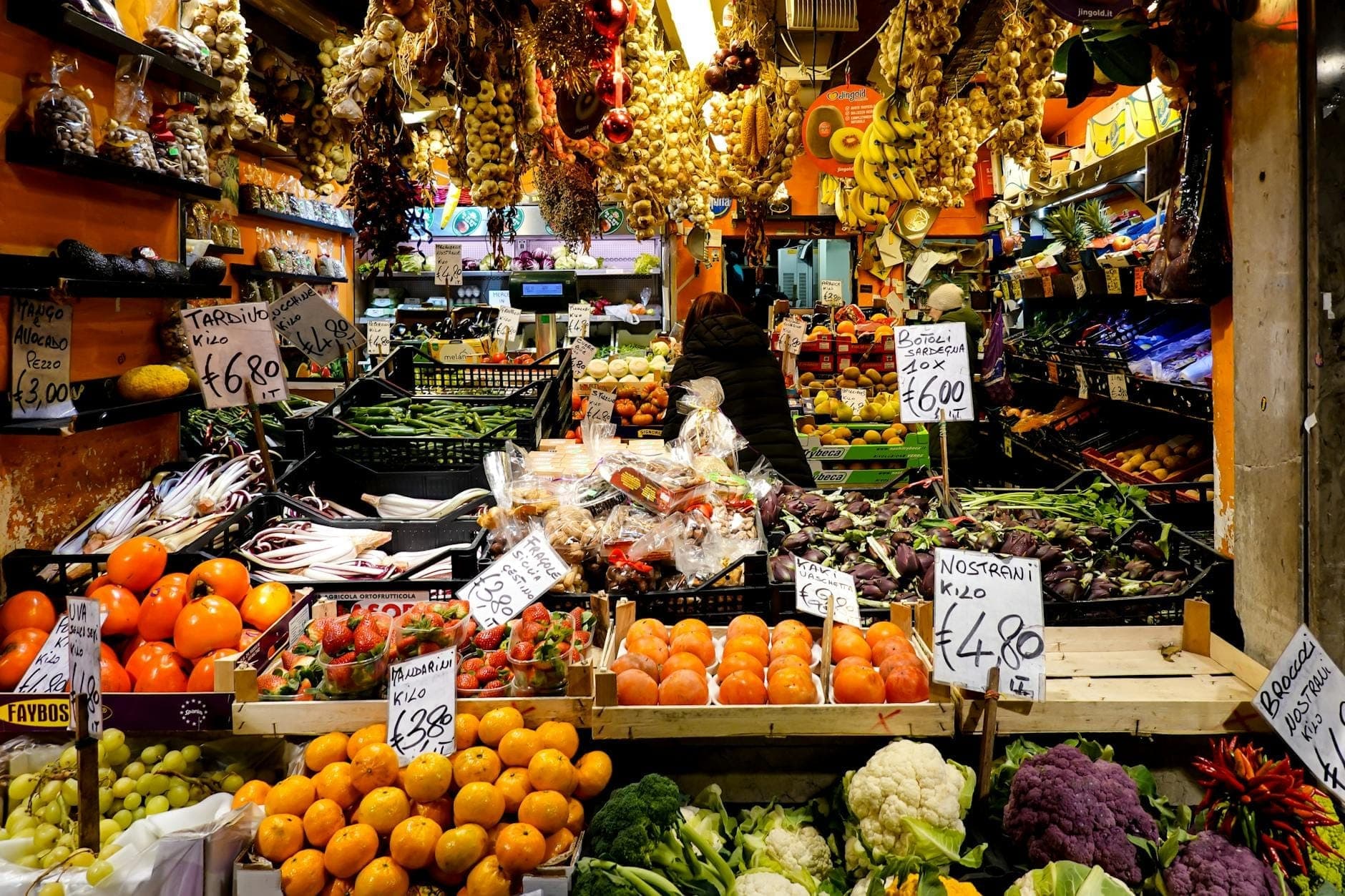 Exposição colorida de frutas e legumes da época com placas de preços numa movimentada banca de mercado romano.