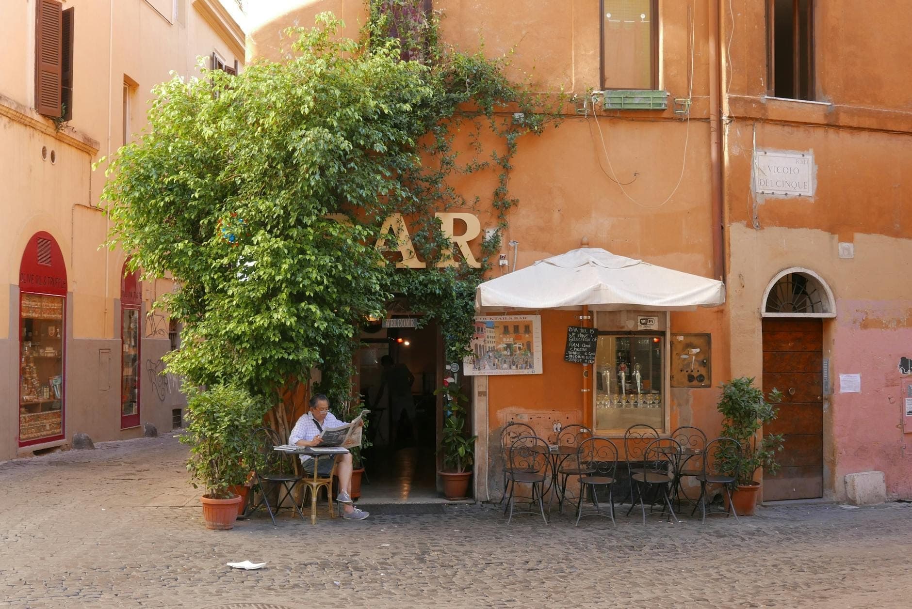 Vista exterior de um charmoso bar romano com assentos numa rua de paralelepípedos, paredes laranjas e uma pessoa a saborear um café numa mesa.