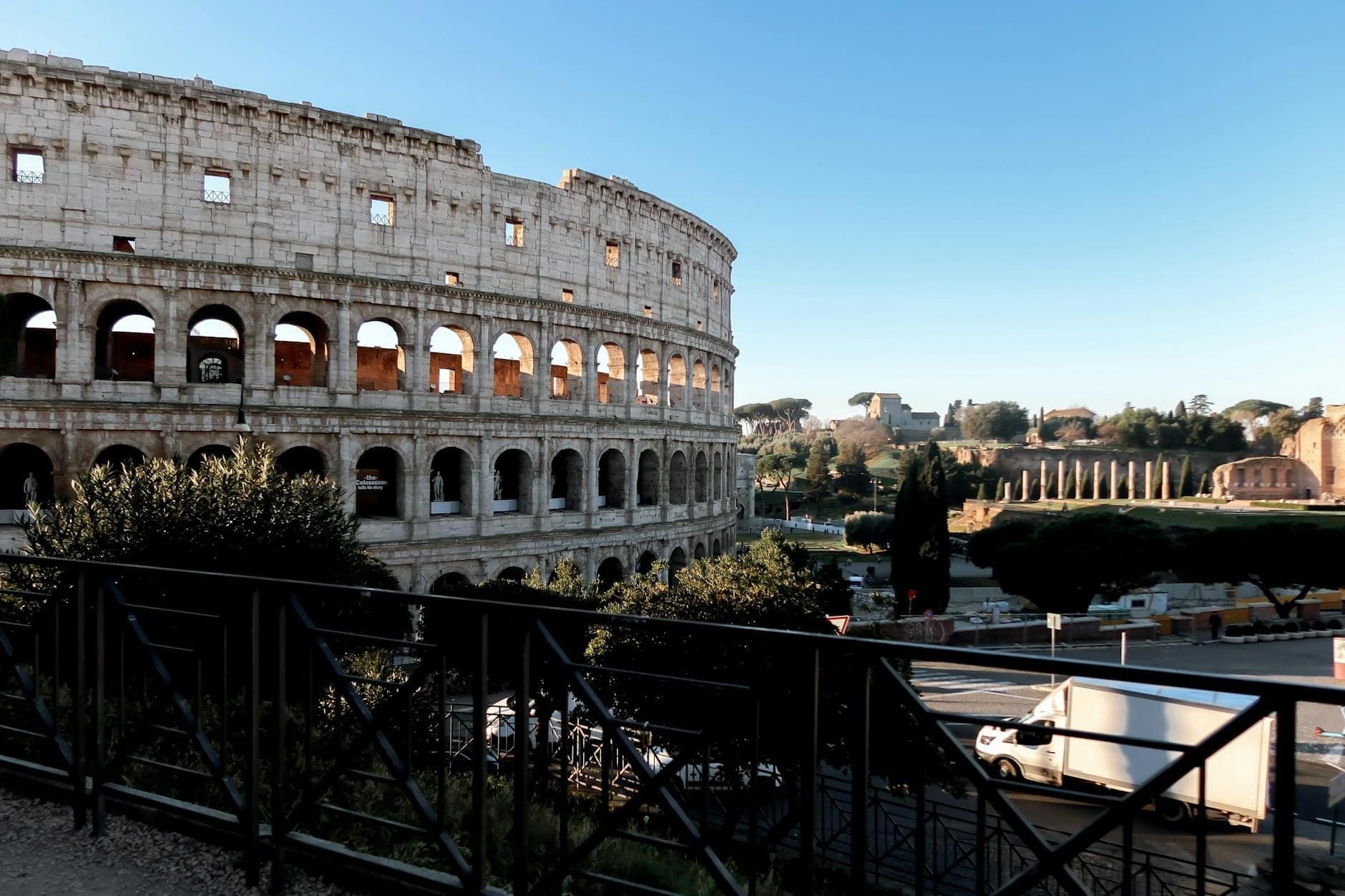 Vue large de jour du Colisée à Rome avec le Forum Romain visible en arrière-plan sous un ciel bleu dégagé.
