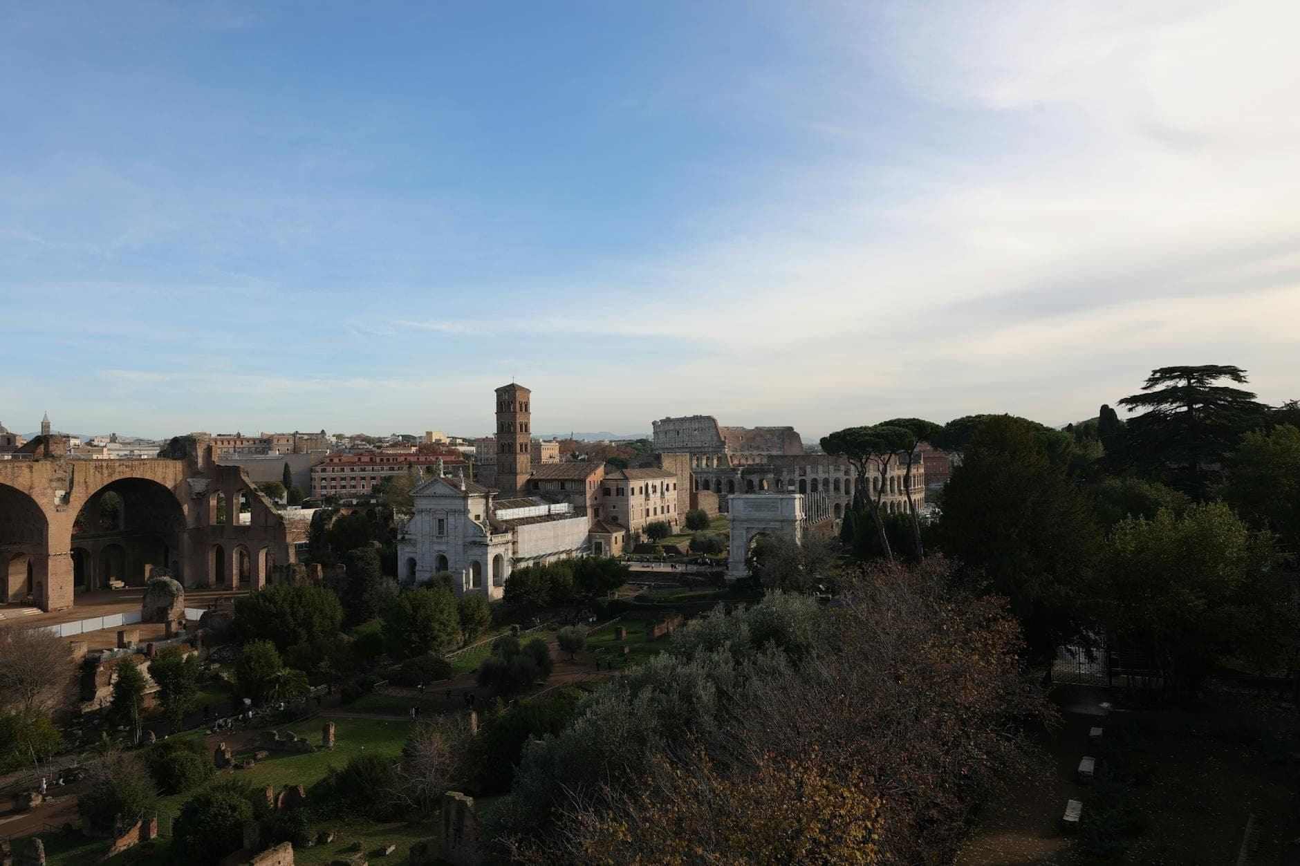 Vue panoramique sur le Forum Romain avec des ruines, de la verdure et un paysage urbain en arrière-plan sous un ciel bleu.