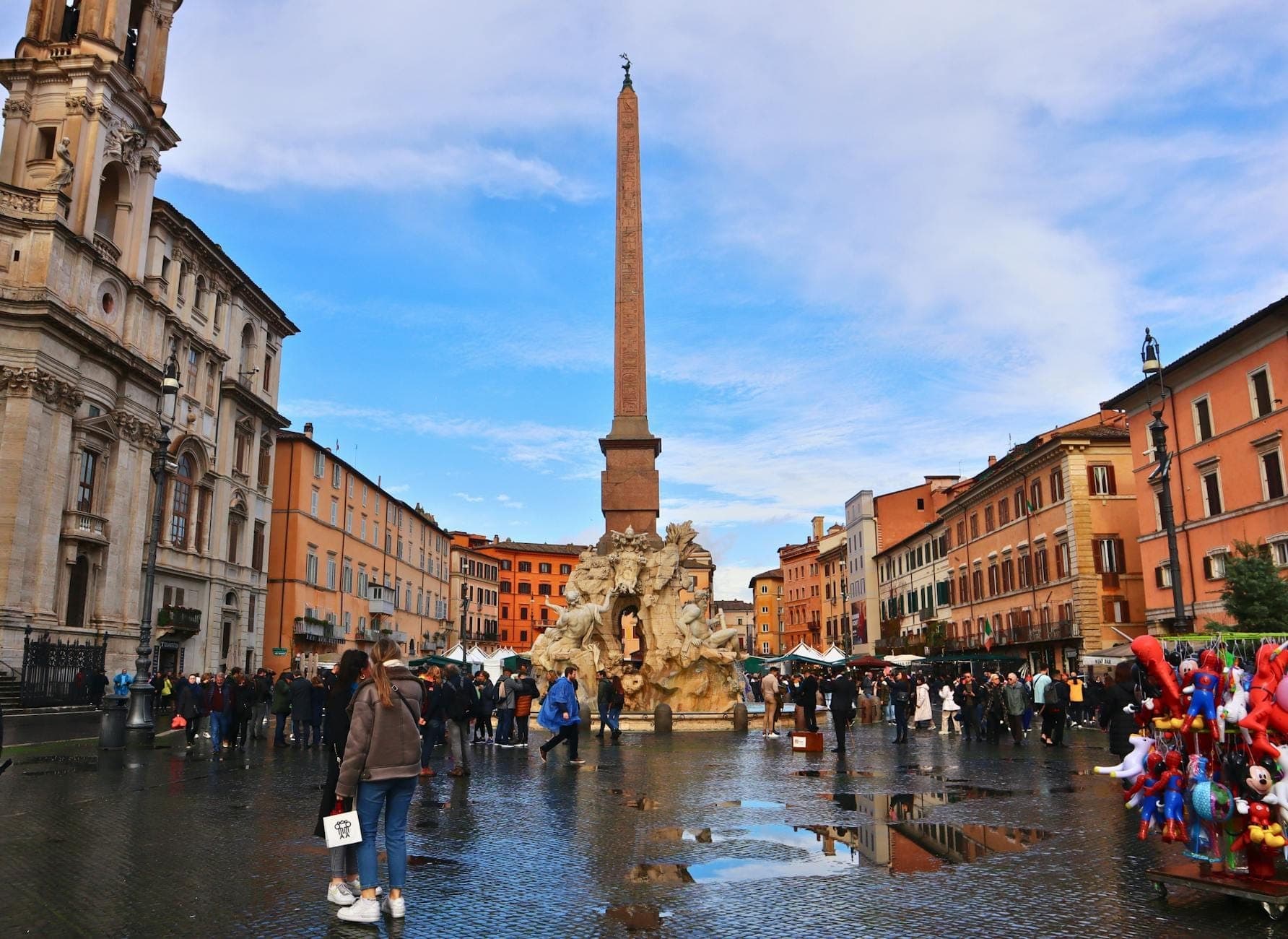 วิวกว้างของ Piazza Navona ในกรุงโรม พร้อมฝูงชน น้ำพุ เสาโอเบลิสก์กลางจัตุรัส อาคารสีสันสดใส และโบสถ์บาโรกภายใต้ท้องฟ้าสีฟ้าสดใส