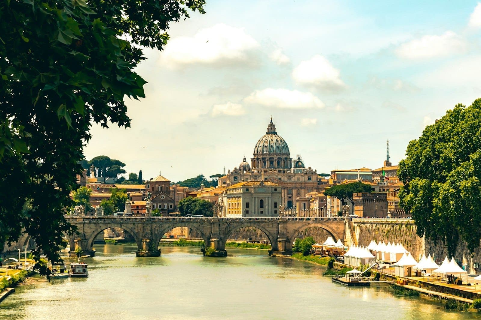 View of Rome featuring the dome of St. Peter's Basilica, the Sant'Angelo Bridge, and the Tiber River framed by trees on a sunny day.