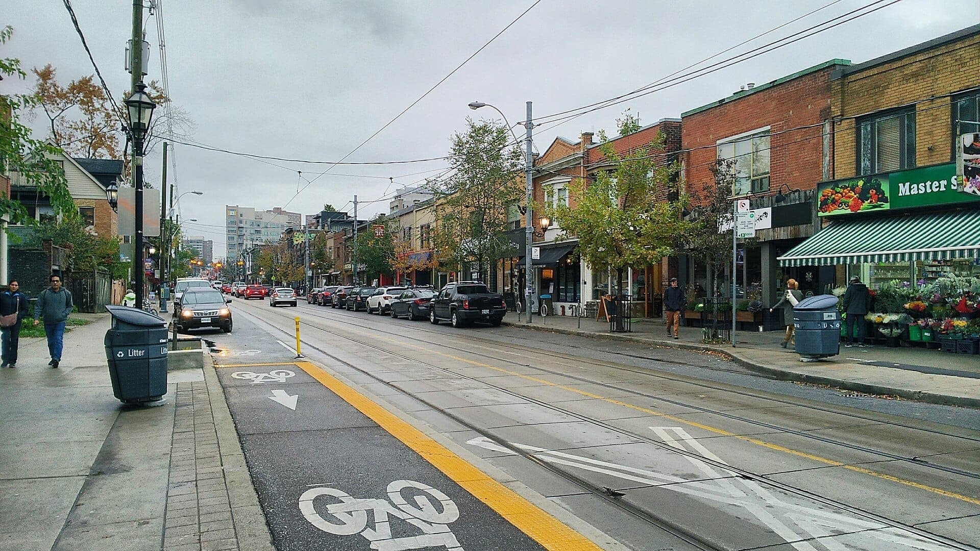 Vista stradale del quartiere Roncesvalles Village a Toronto in una giornata nuvolosa, con negozi indipendenti, auto parcheggiate, piste ciclabili e pedoni.