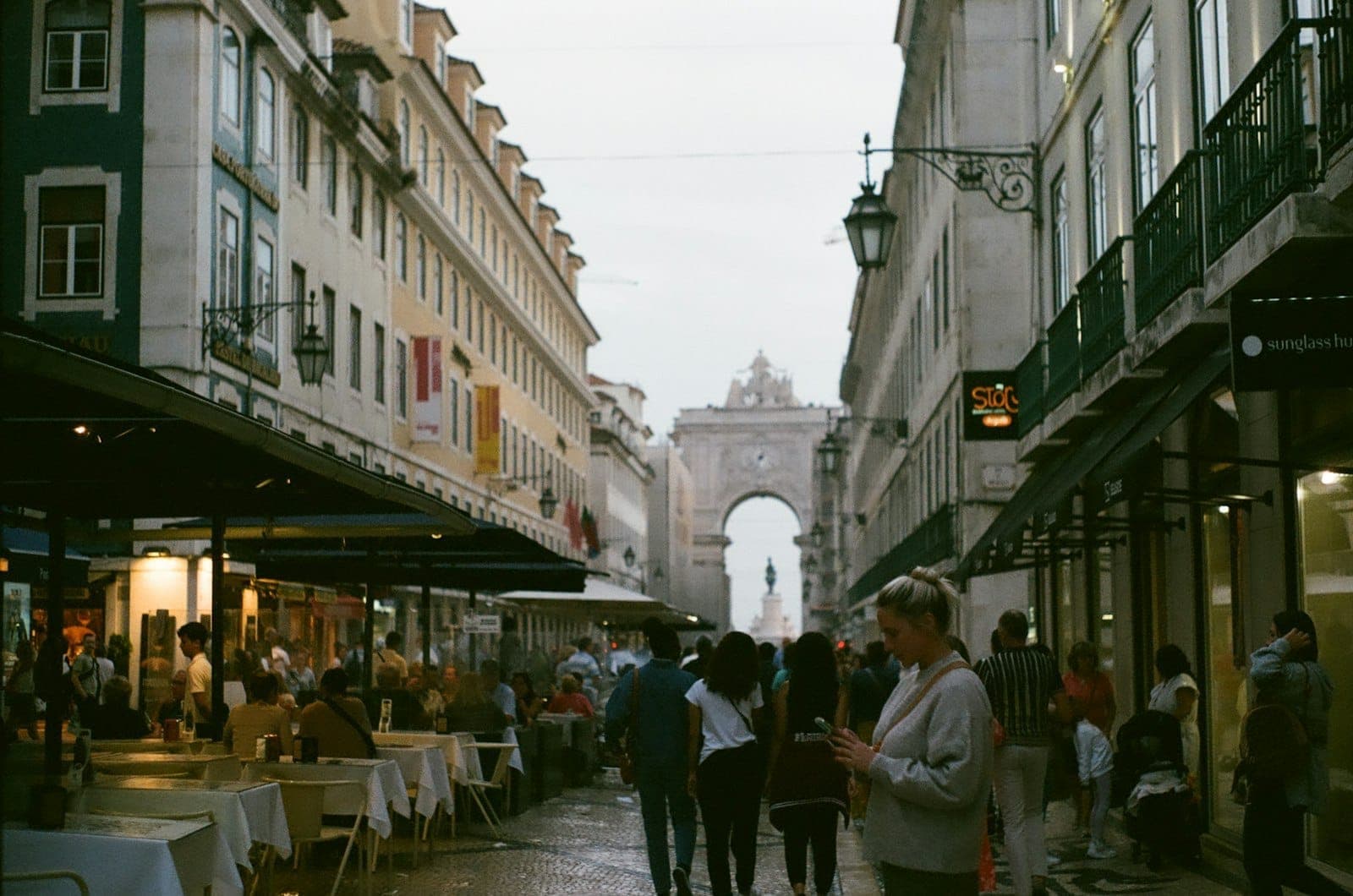 Pedestres caminhando pela Rua Augusta, uma rua de paralelepípedos ladeada de cafés e lojas, com o arco triunfal ao fundo.