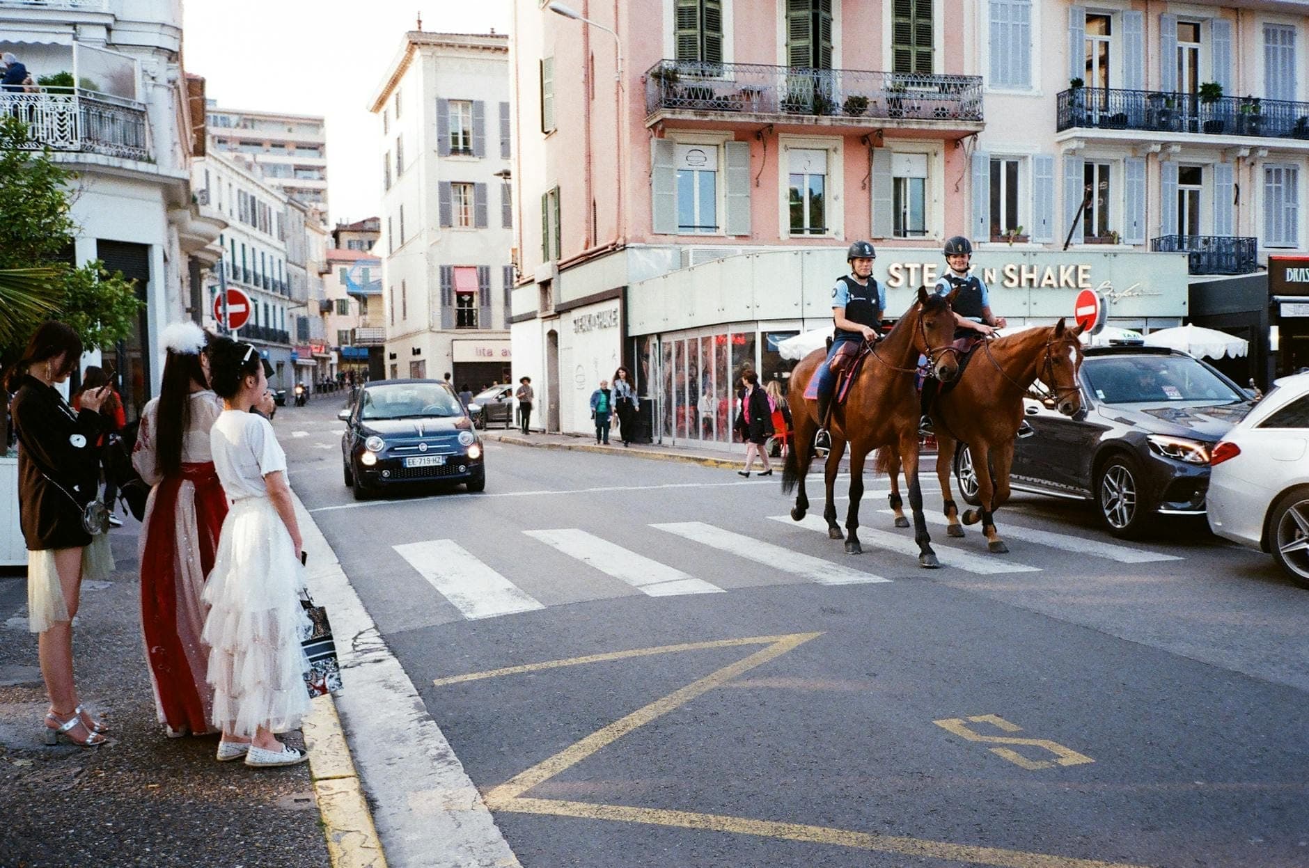 Rue d'Antibes в Каннах: здания в пастельных тонах, люди в костюмах, проезжающие автомобили и конная полиция, патрулирующая оживлённую улицу.
