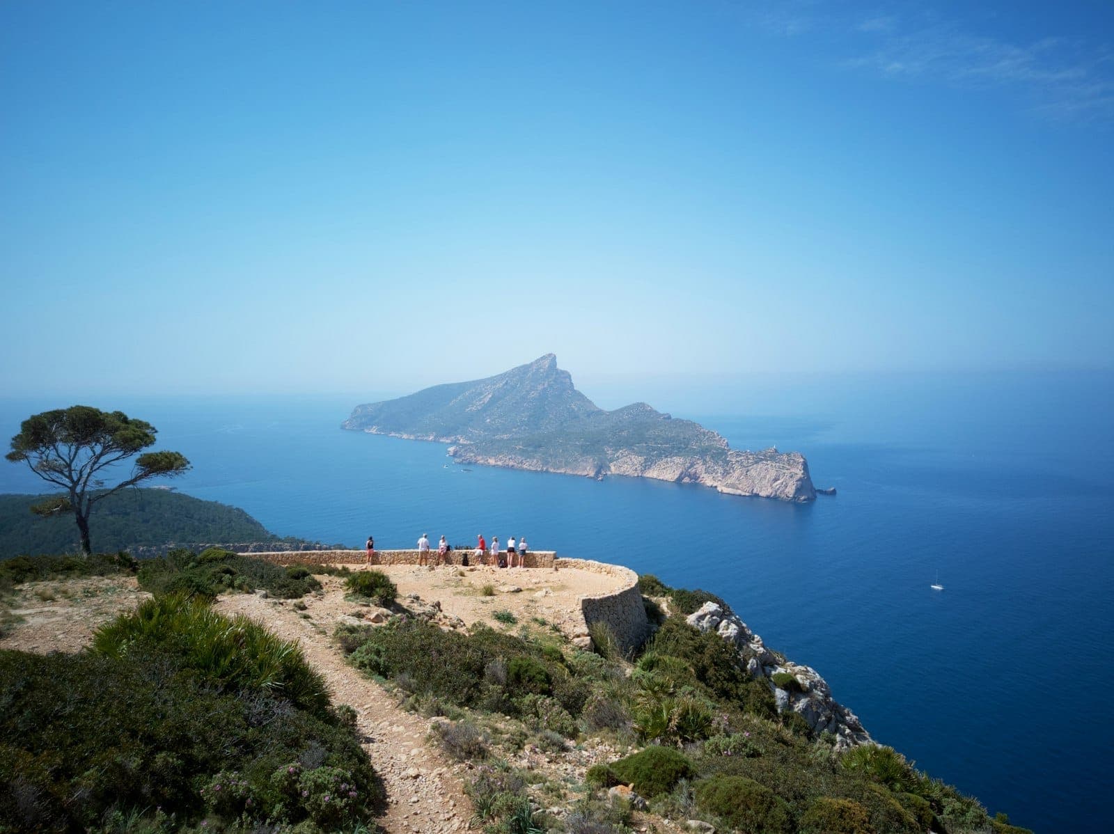 Blick auf die Insel Sa Dragonera von einem Aussichtspunkt auf einer Klippe, mit zerklüftetem Gelände, blauem Meer und Besuchern, die die Naturlandschaft genießen.