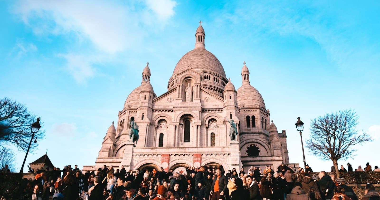 Une foule de visiteurs se presse sur les marches menant à la basilique du Sacré-Cœur de Montmartre, sous un ciel d'un bleu éclatant, avec ses célèbres dômes en arrière-plan.
