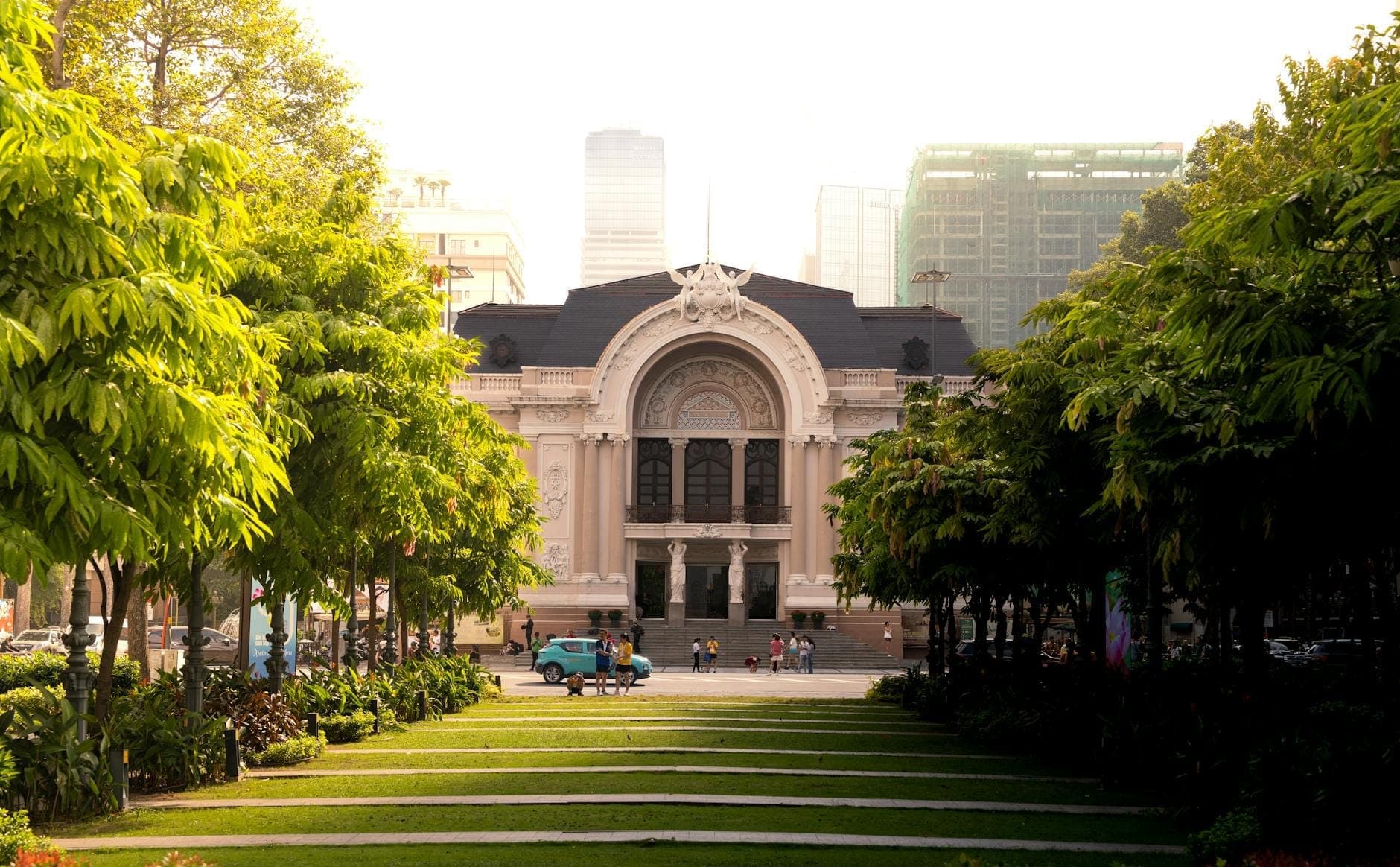 Ang maringal na Saigon Opera House na napalibutan ng malalabong puno at isang maliwanag na landas ng hardin, na may mga modernong gusali ng lungsod sa likod, na lumilikha ng isang masayang eksena ng pagbiyahe sa Ho Chi Minh City.