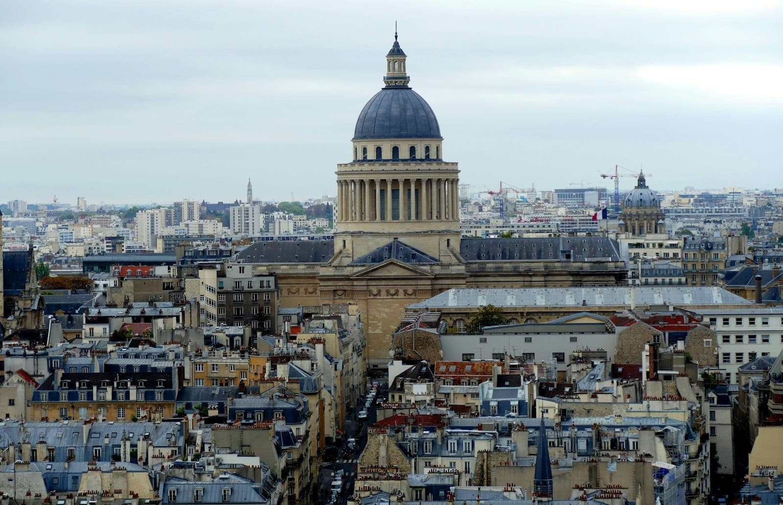 A panoramic view of the Pantheon in the Latin Quarter, with classic Parisian rooftops and cityscape stretching into the distance under a cloudy sky.