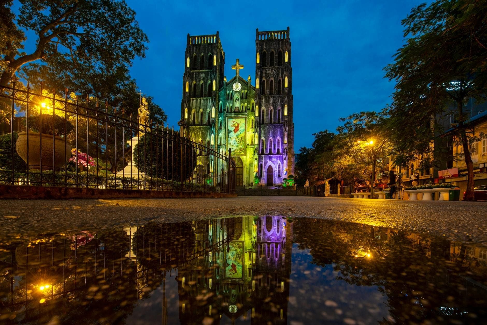 Katedra św. Józefa w Hanoi rozświetlona o zmierzchu – bliźniacze wieże i gotycka fasada odbijają się w kałuży, a całość otaczają drzewa i ożywiony plac.