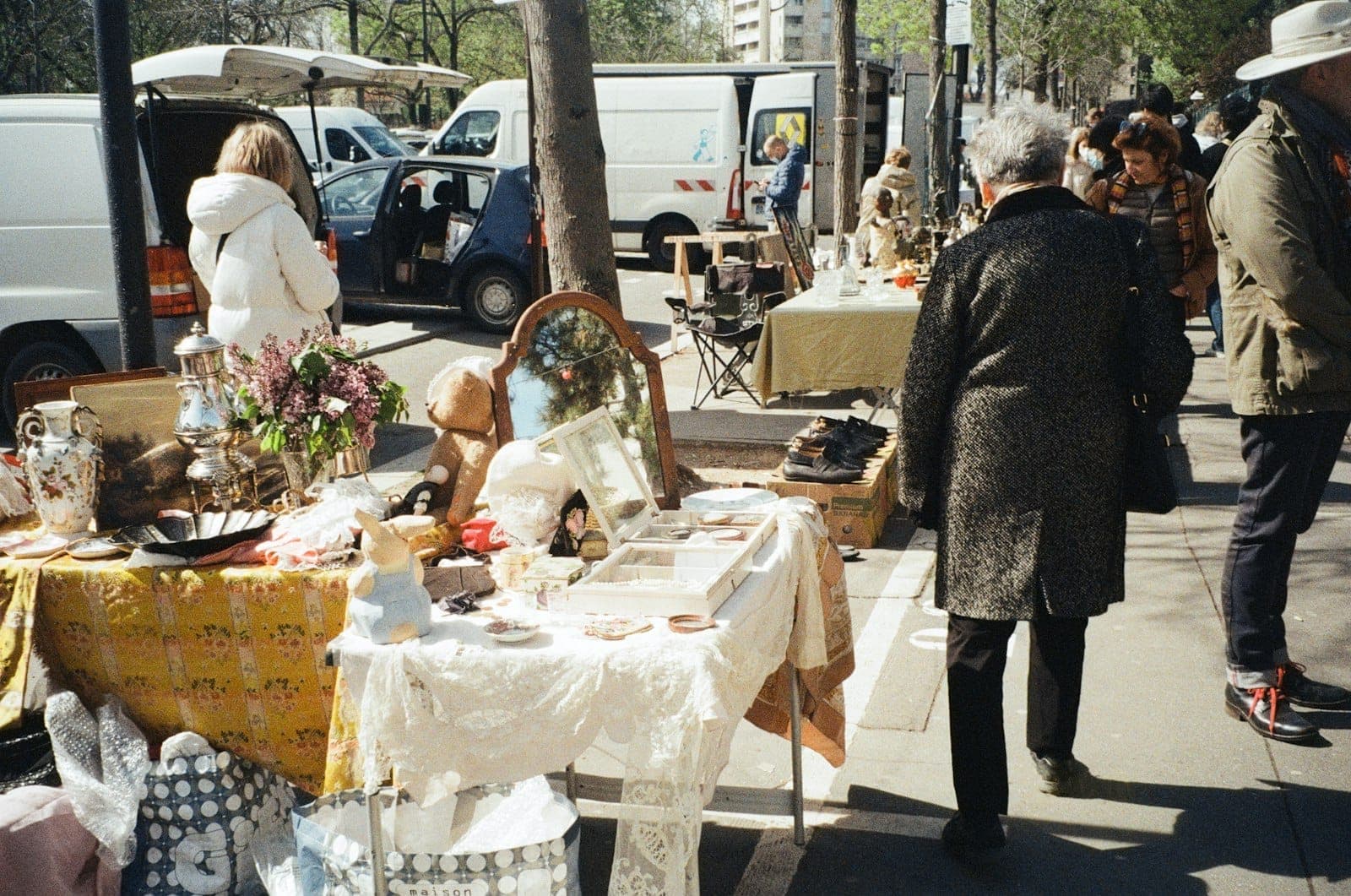Outdoor flea market scene with people browsing tables filled with antiques, vintage items, and collectibles along a Paris street on a sunny day.