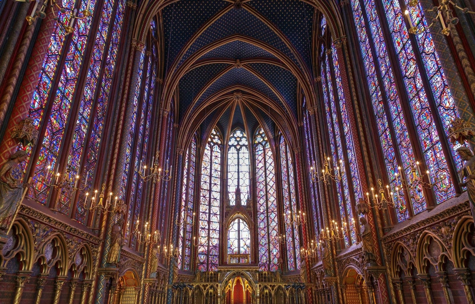Interior view of Sainte-Chapelle’s upper chapel, showing soaring stained glass walls, vaulted ceiling, glowing chandeliers, and intricate Gothic stonework bathed in colored light.