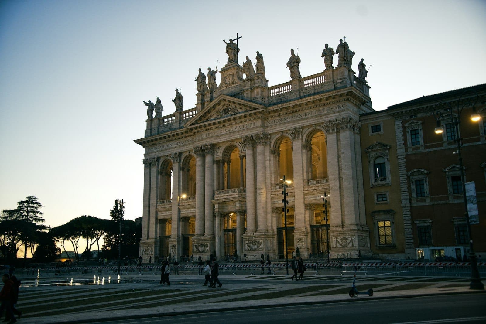 Oświetlona fasada bazyliki San Giovanni in Laterano o zmierzchu, ze statuami wzdłuż dachu i ludźmi spacerującymi na pierwszym planie.