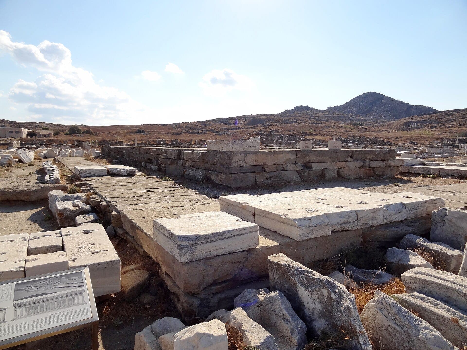 Antike Steinruinen des Heiligtums des Apollon auf Delos, Griechenland, umgeben von felsiger Landschaft und klarem blauem Himmel.