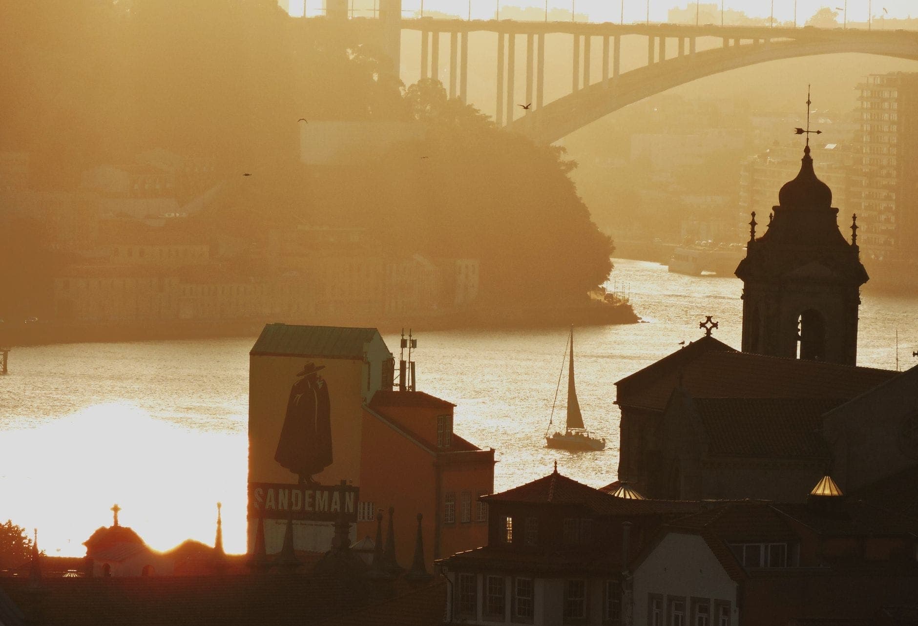 Vue dorée au coucher du soleil sur le Douro avec l'enseigne des caves Sandeman, des barques et le pont iconique de Porto en arrière-plan.