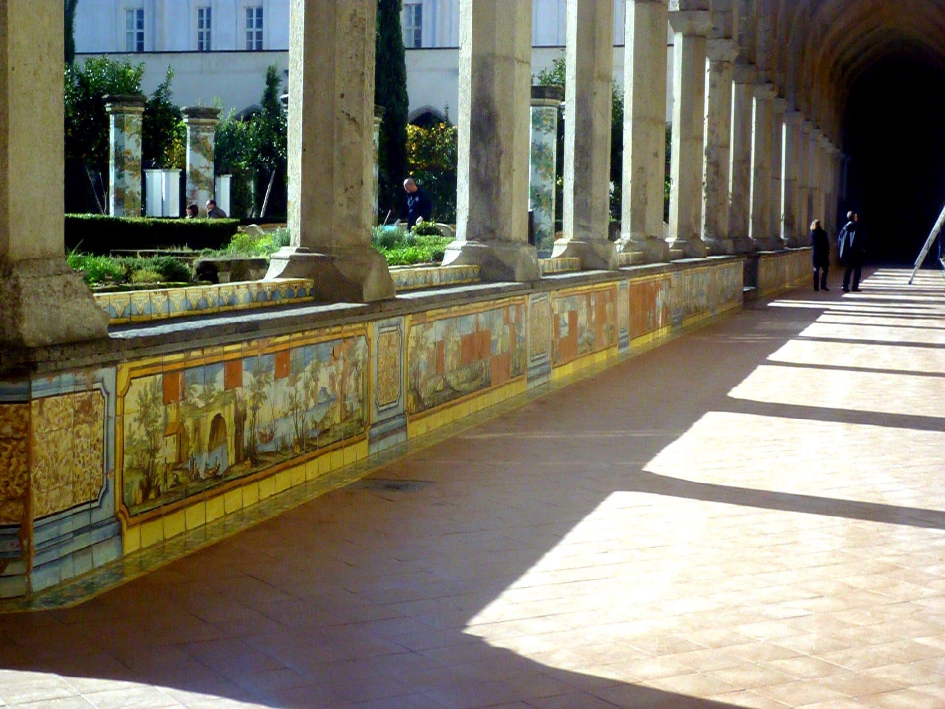 Paredes coloridas com azulejos de majólica e colunas de pedra ladeiam o tranquilo corredor do claustro do Complesso Monumentale di Santa Chiara em Nápoles sob um sol radiante.
