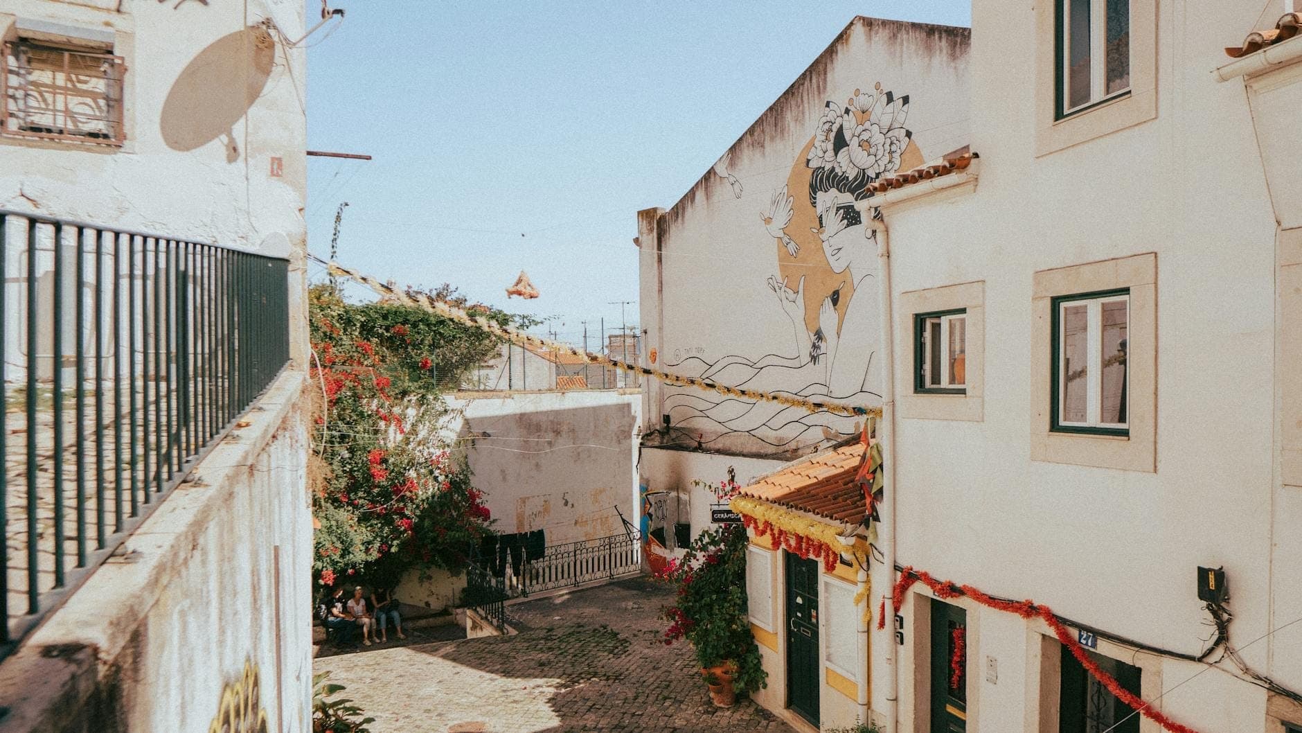 Bairro iluminado pelo sol em Lisboa com cordas de luzes, mural de arte de rua e algumas pessoas sentadas numa pequena praça, evocando um ambiente festivo.